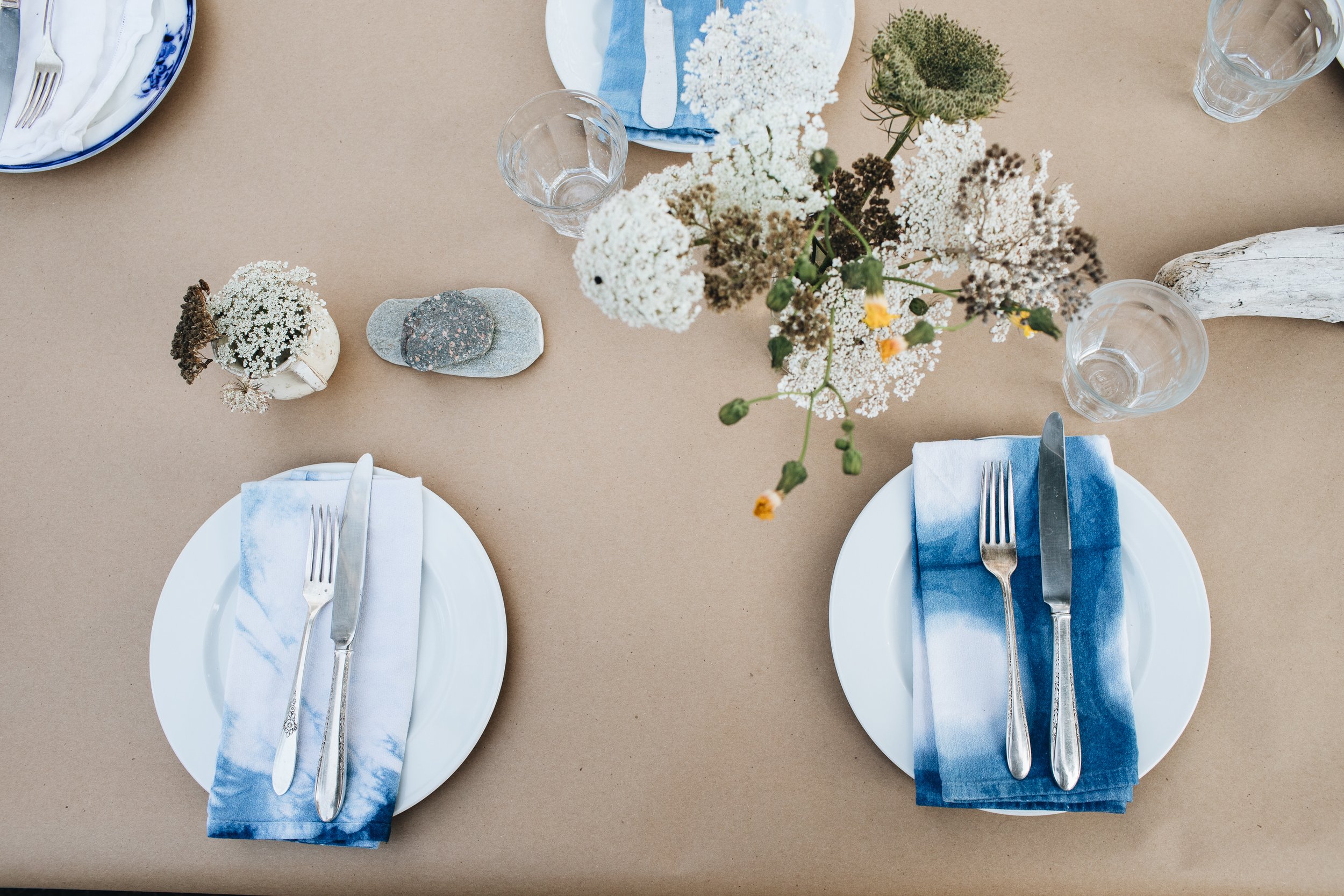 Table setting with white plates, blue and white napkins, forks, knives, clear glasses, and a floral centerpiece with white, green, and yellow flowers, on beige tablecloth.