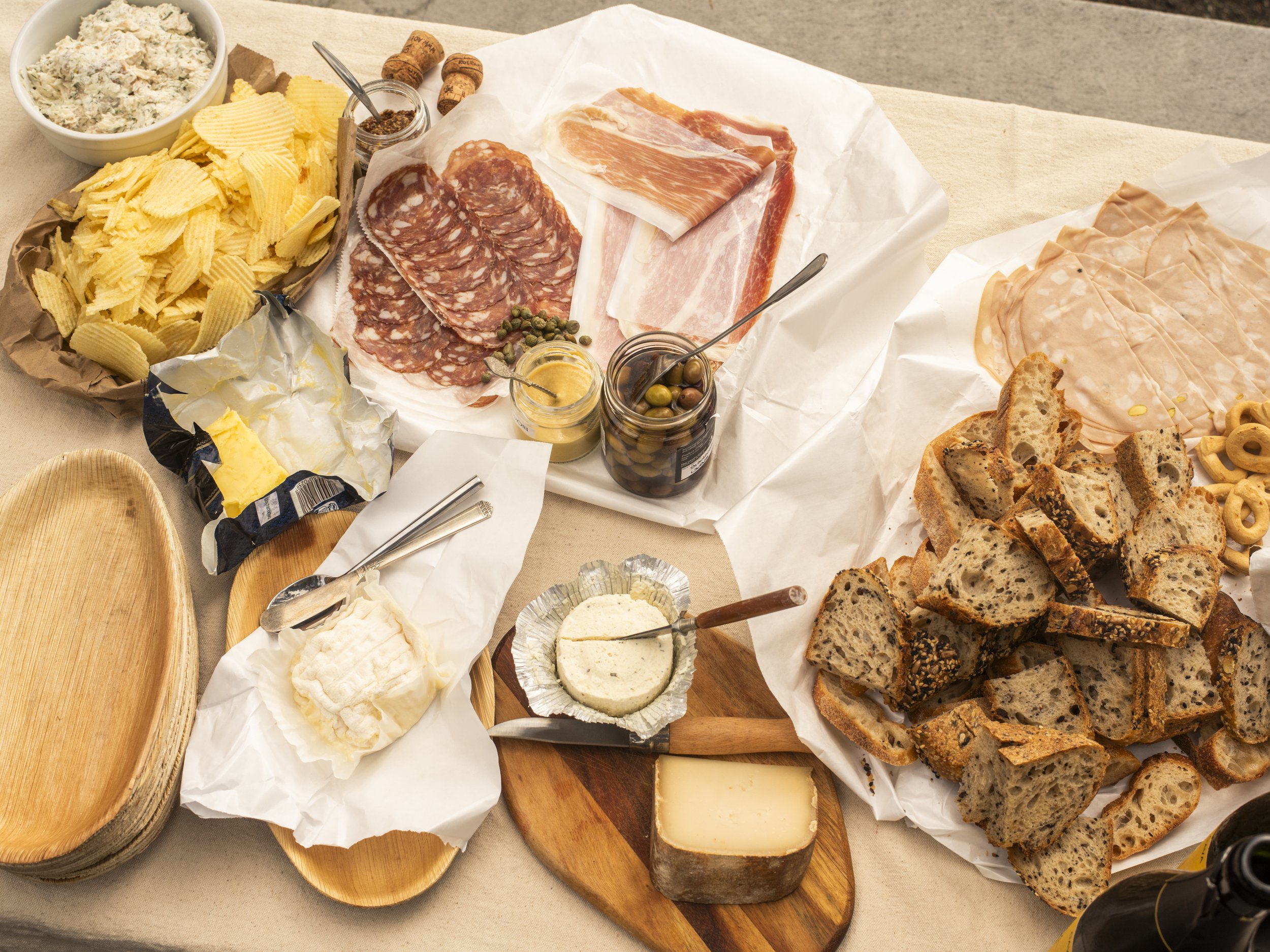 A spread of charcuterie board with assorted meats, cheeses, bread, chips, olives, and condiments.