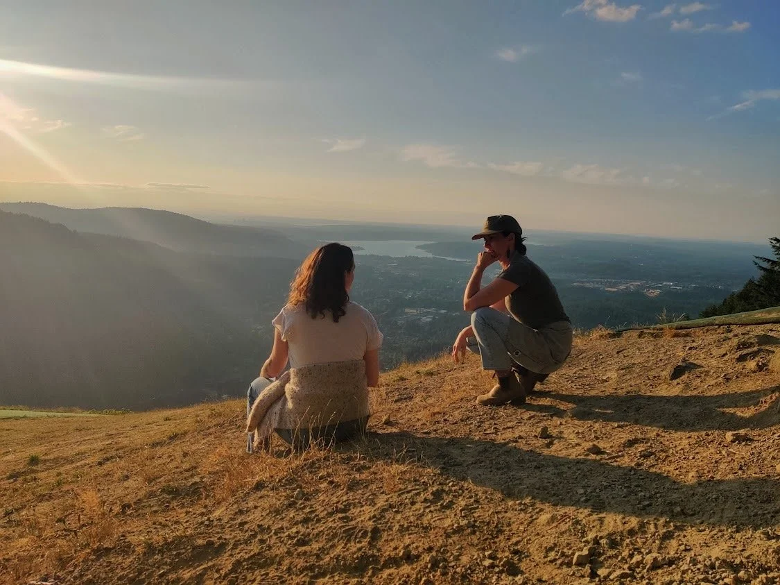 Two women sitting and squatting on a hilltop overlooking a scenic landscape at sunset, with a lake and mountains in the distance.