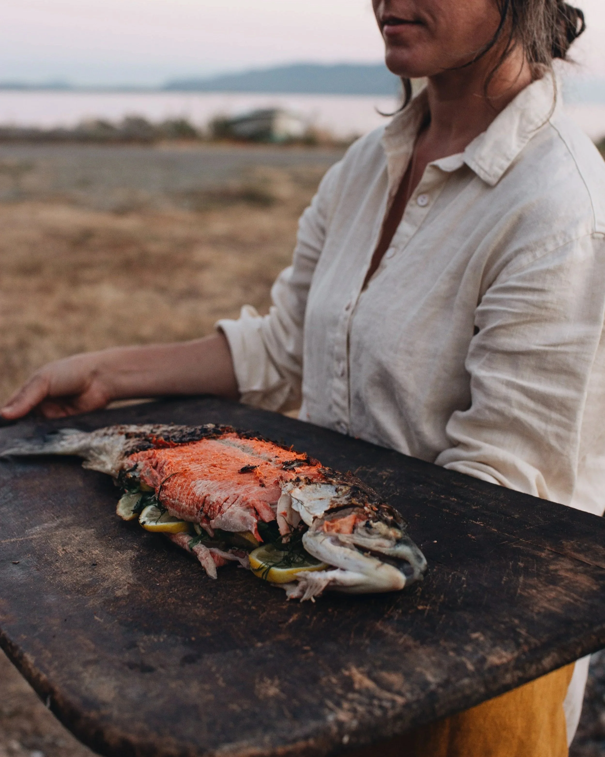 A person holding a large, cooked fish on a wooden cutting board outdoors near a body of water, with a landscape of mountains and sky in the background.