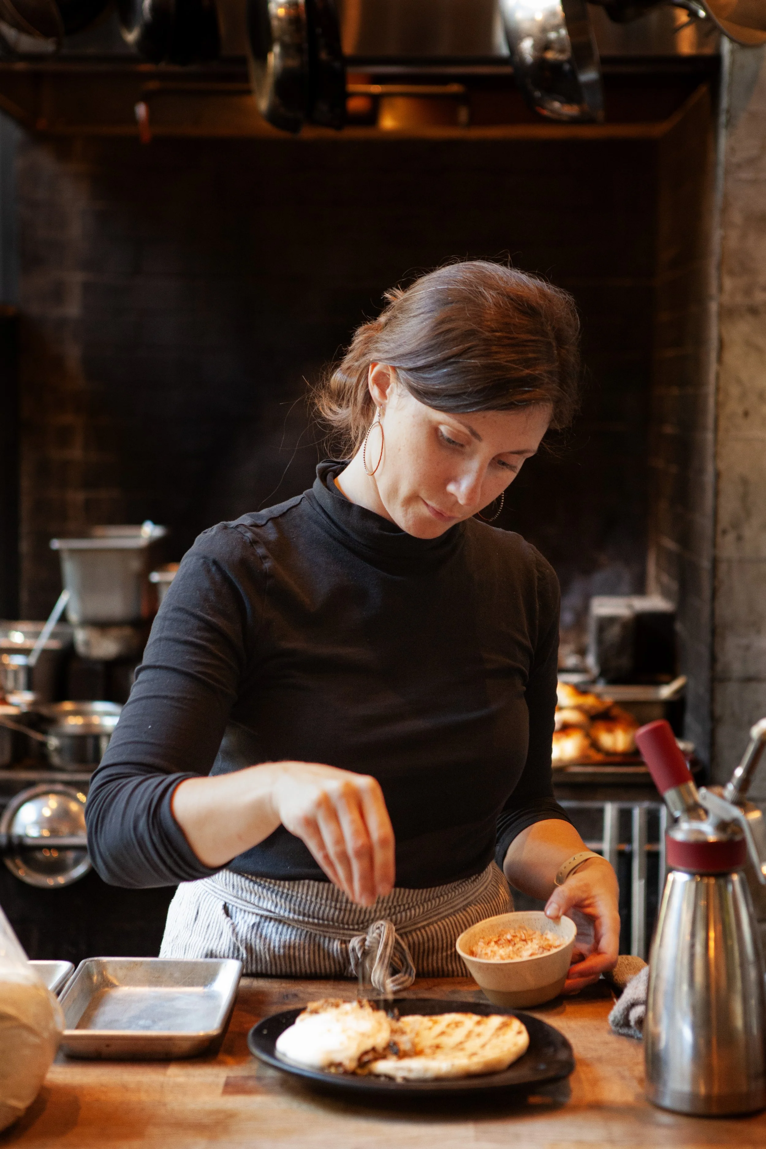 A woman in a black top and striped apron preparing food in a kitchen with a brick wall and a fireplace in the background.
