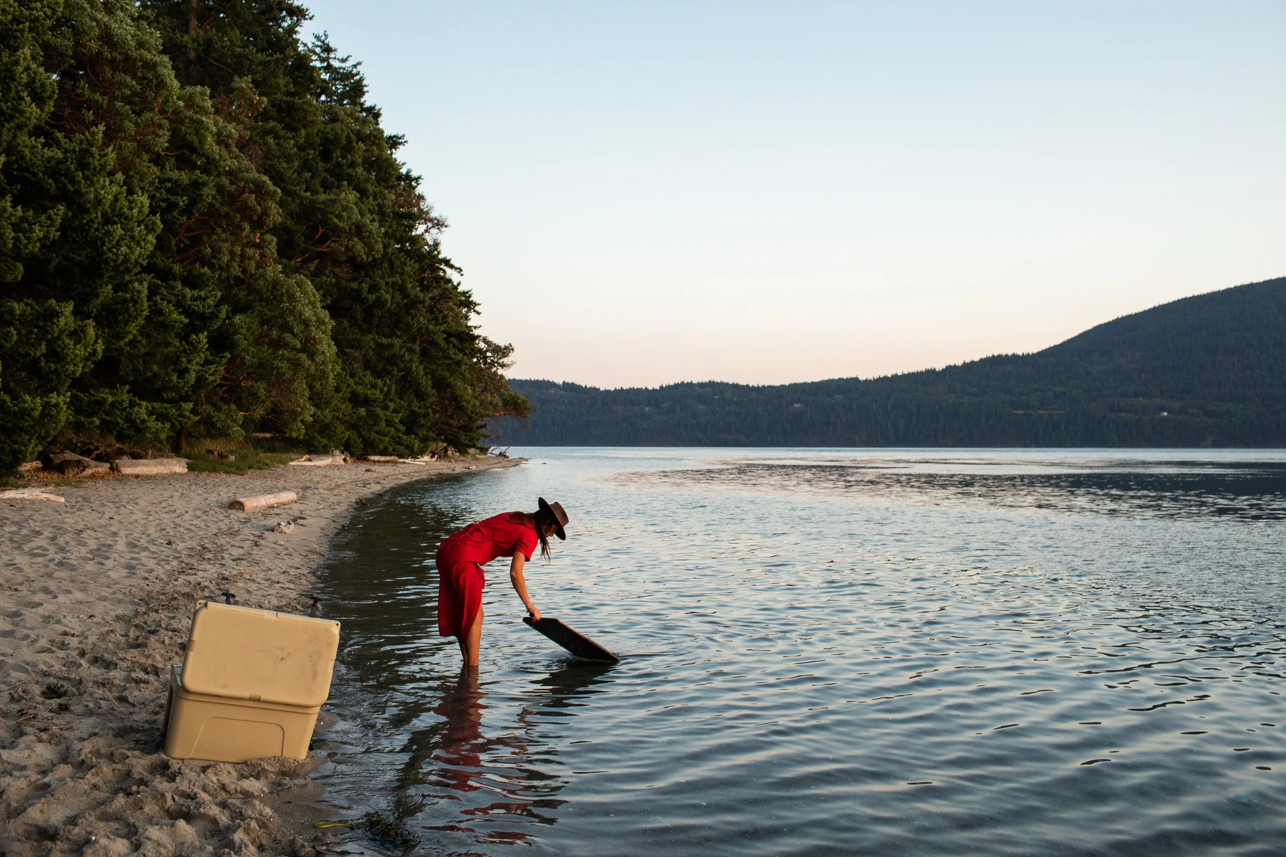 A woman in a red dress and wide-brimmed hat stands in shallow water at the edge of a sandy beach, holding a paddleboard. There is a large beige cooler on the beach beside her. The backdrop features a forested shoreline and distant wooded hills under 