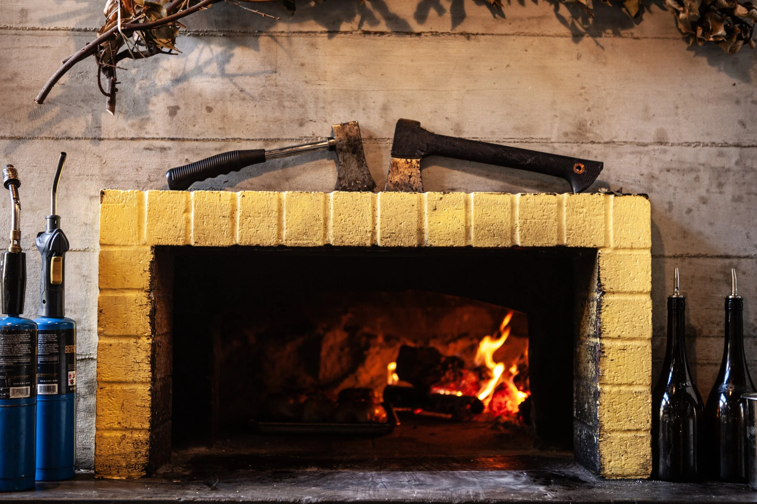 A fireplace with burning logs, two axes on the mantel, and various tools and bottles nearby.