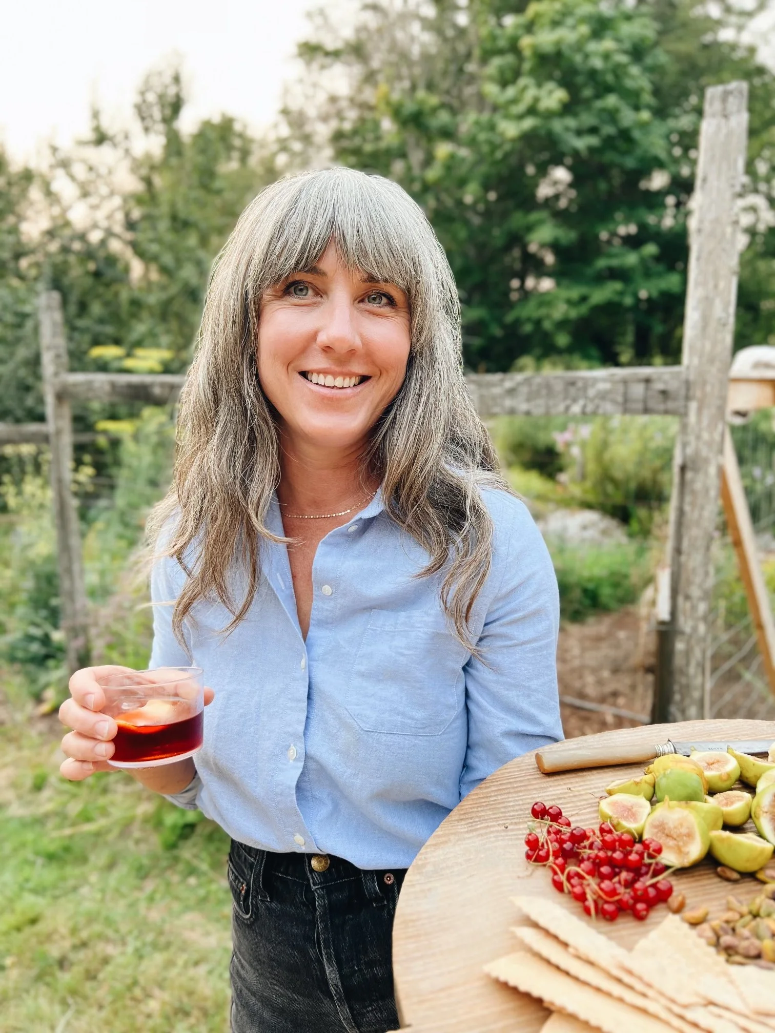 A woman with long gray hair wearing a light blue shirt is holding a glass of red wine outside. There is a wooden table with sliced figs, red berries, crackers, and nuts in front of her. The background shows a garden with trees and a wooden fence.