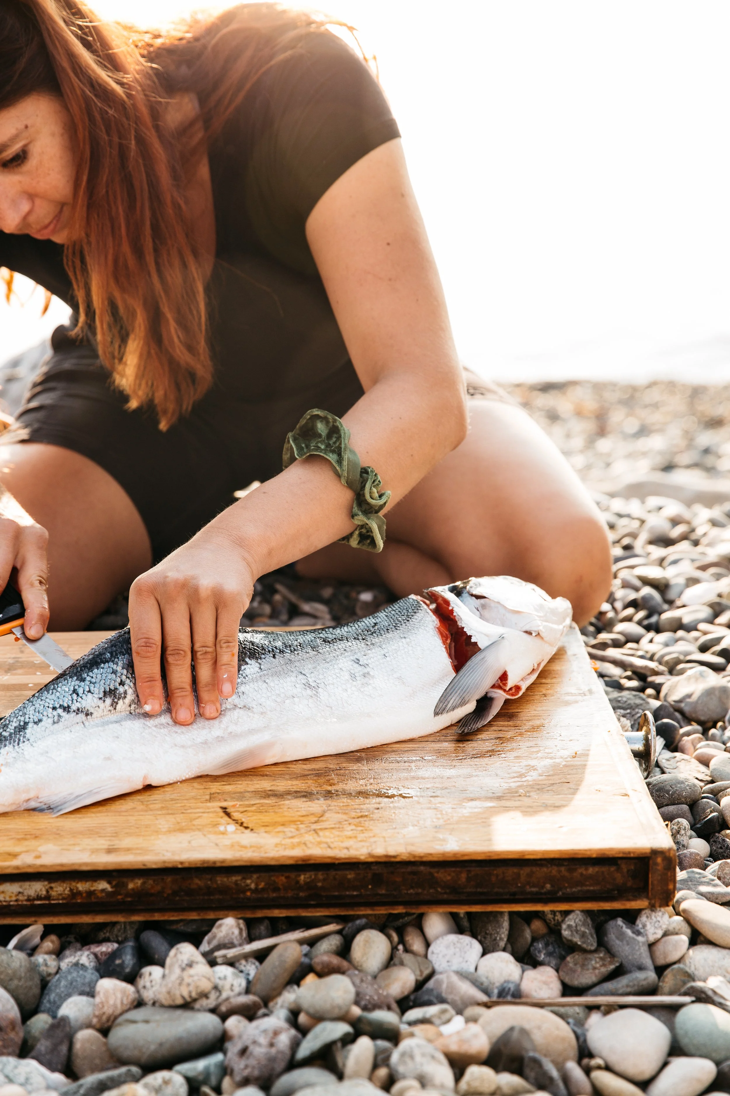 A woman in black clothing preparing a fish on a wooden cutting board on a rocky beach at sunset.