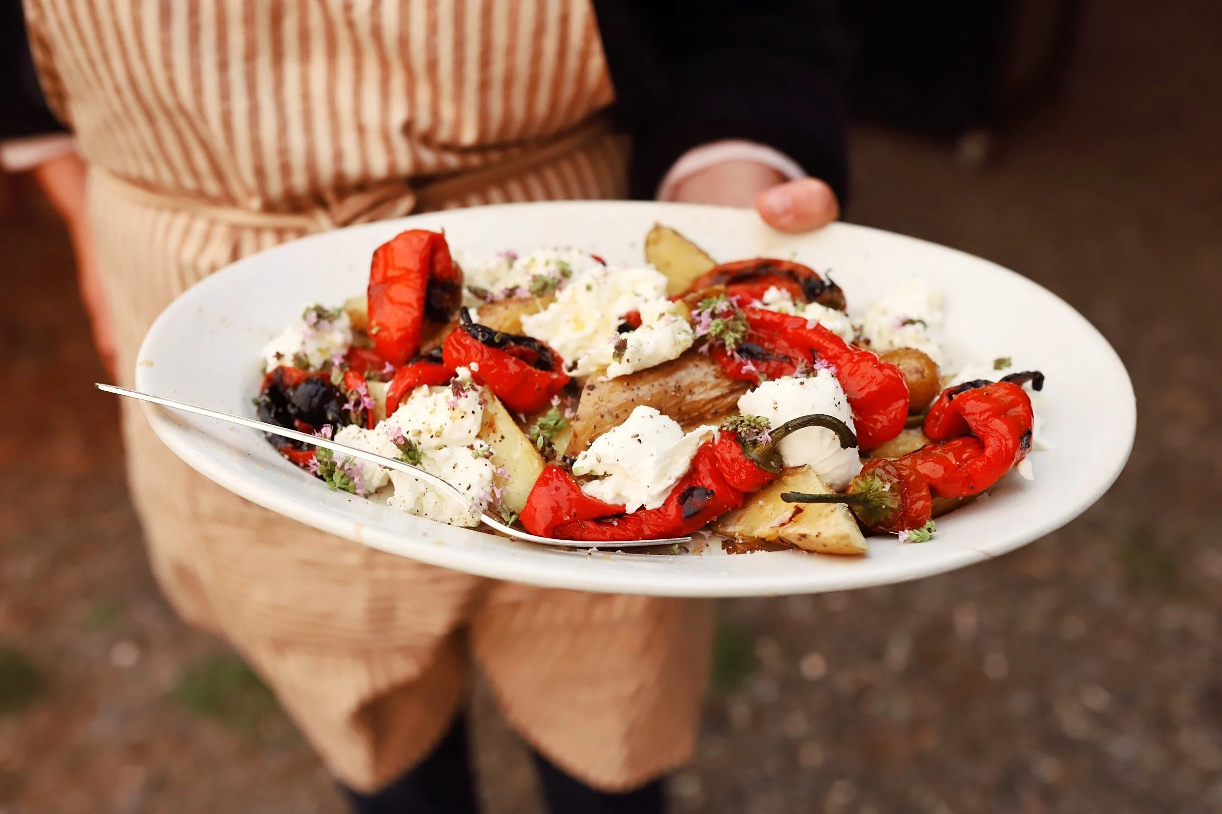 Person holding a white plate with grilled red peppers, potatoes, cheese, and herbs, outdoors.