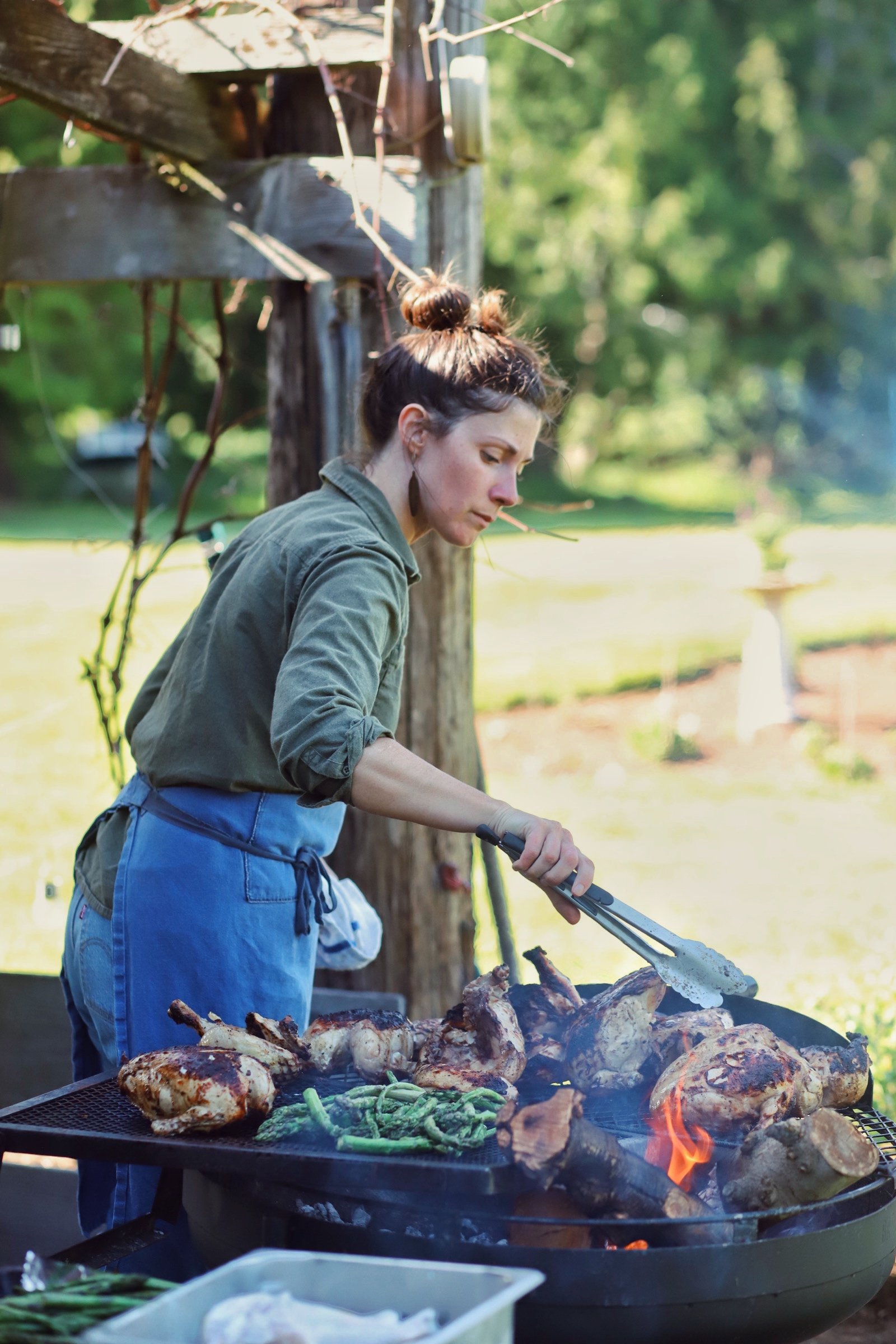 Woman grilling assorted meats and vegetables outdoors on a barbecue grill with green trees in the background.