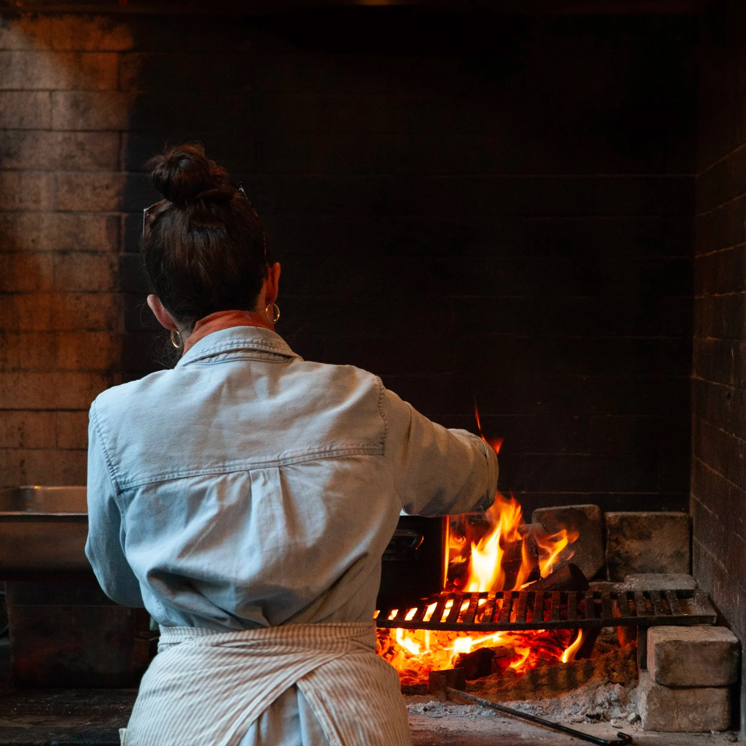 A person cooking near an open brick fireplace with flames visible.