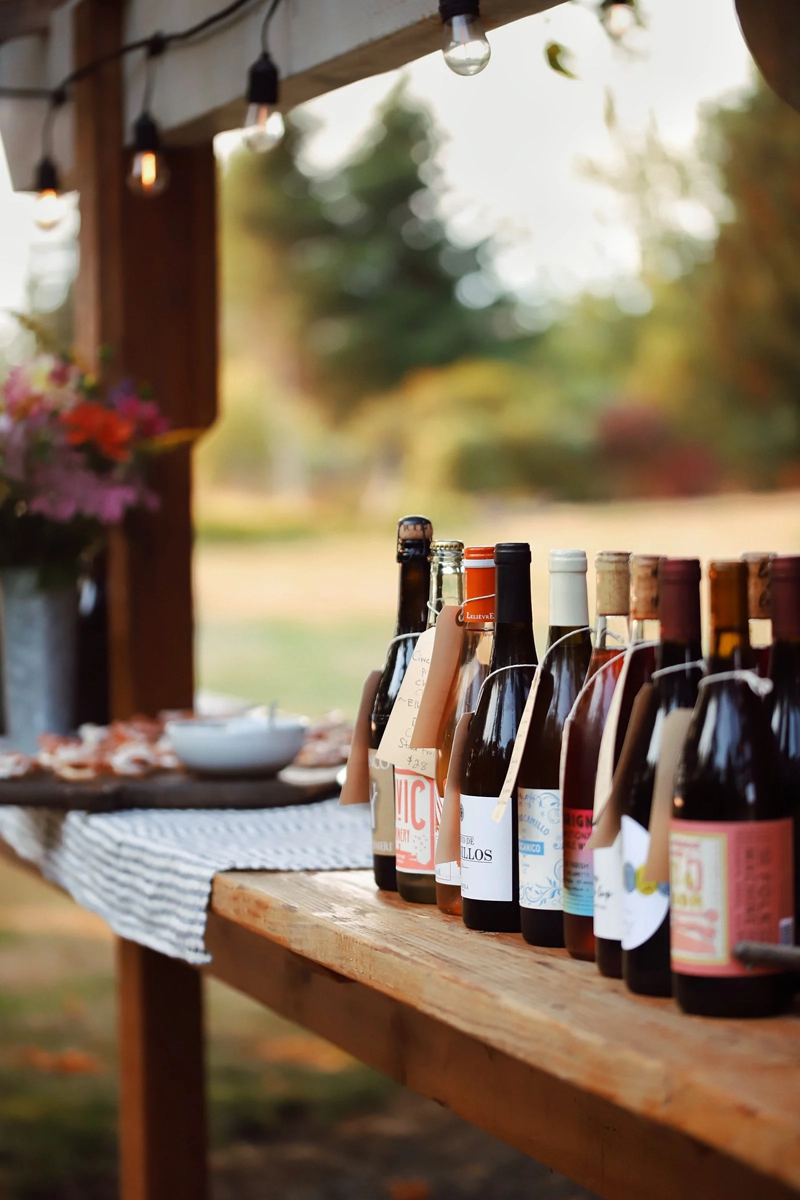 A row of assorted bottles of wine on a wooden table outdoors, with blurred background and string lights overhead.
