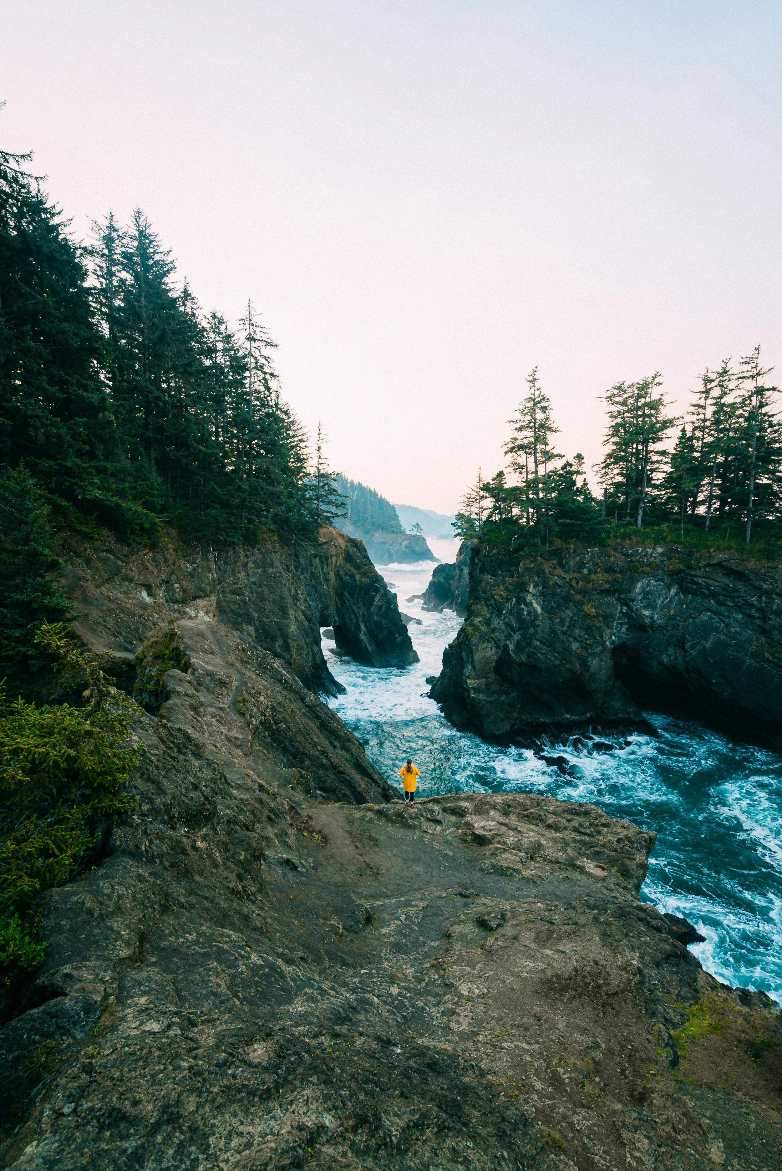A person in a yellow jacket stands on rocky ground overlooking the ocean between two cliffs with trees, with waves crashing below, during sunset.