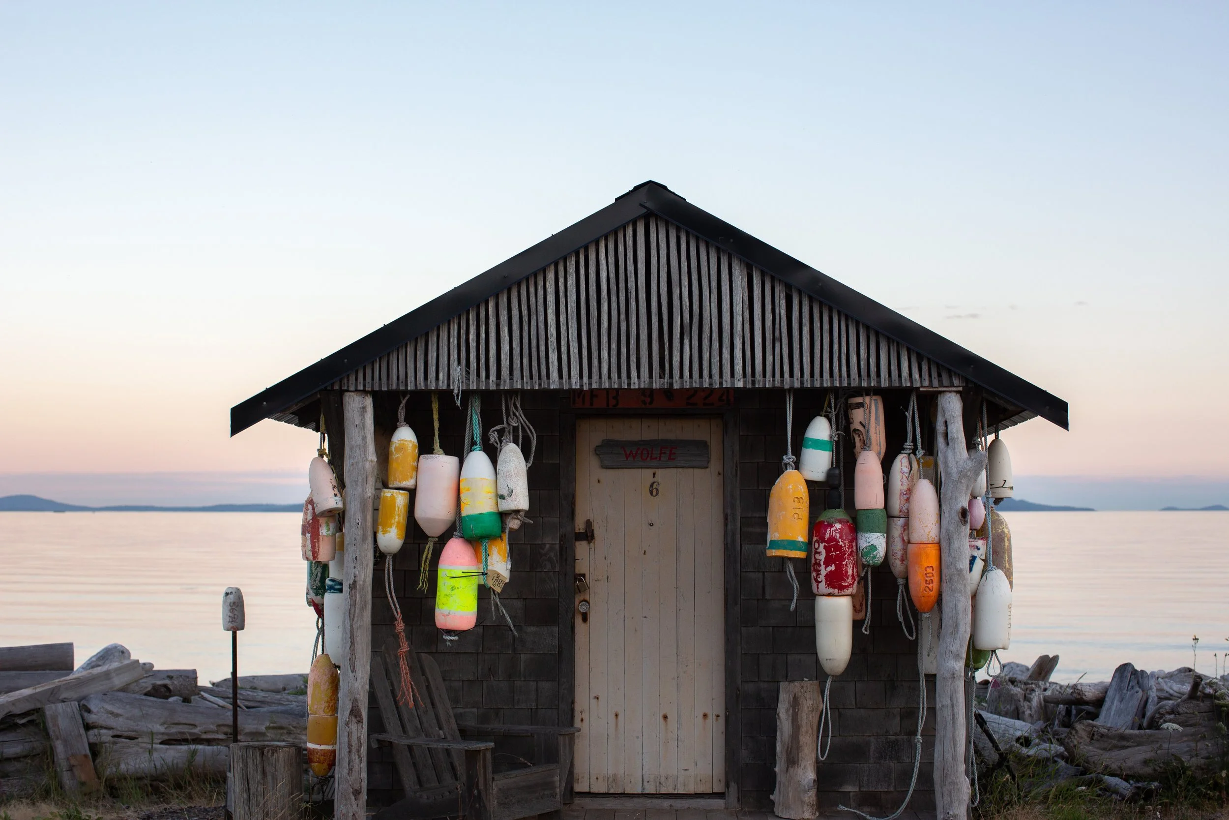 A small wooden boathouse with buoys hanging from its posts, located by a calm body of water during sunset.