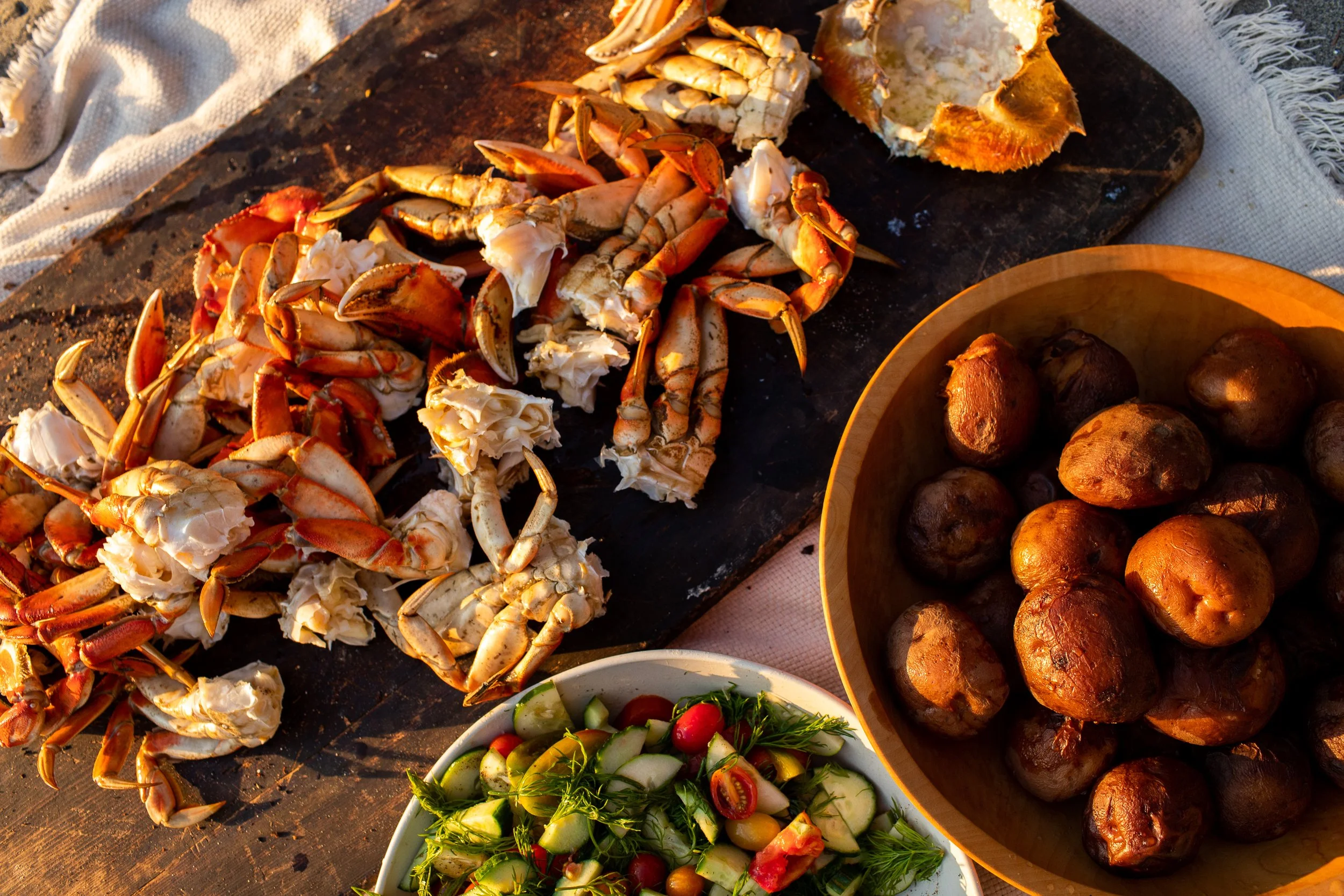 Crabs in pieces on a cutting board, a bowl of cooked potatoes, and a colorful vegetable salad with cucumbers, tomatoes, and herbs, all placed on a rustic wooden surface.