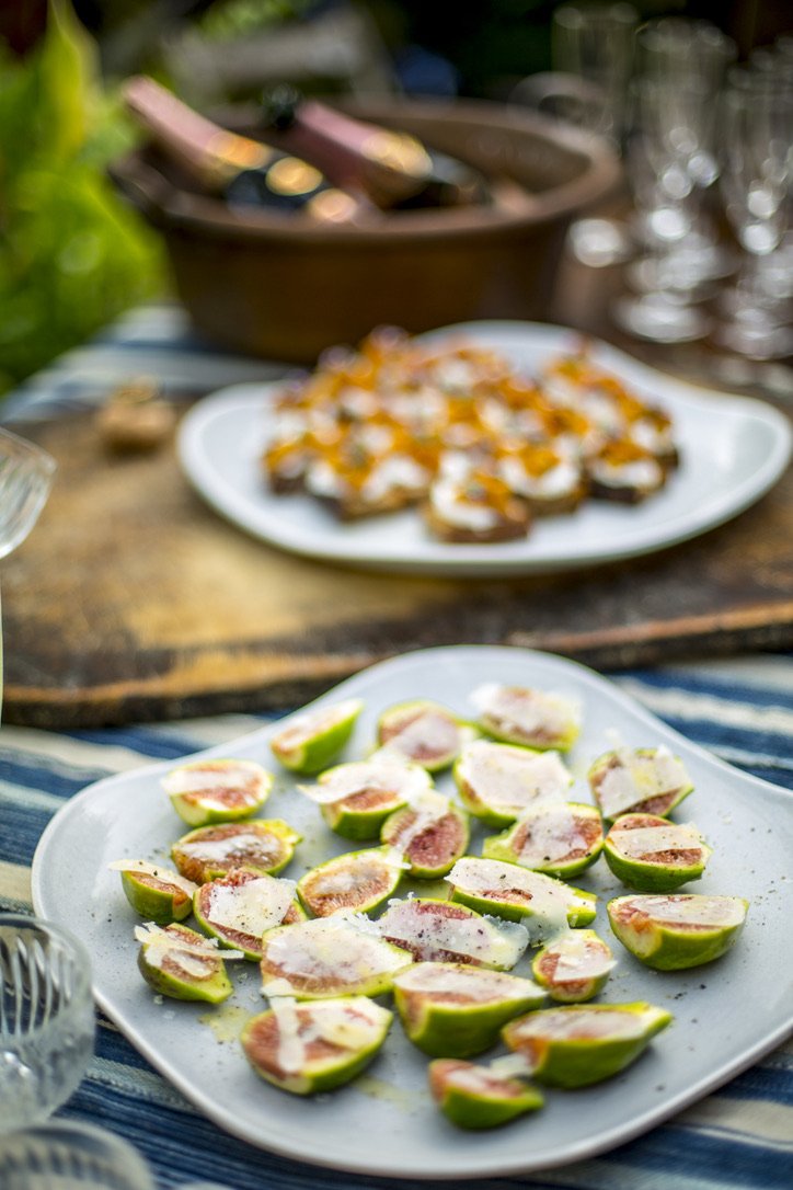 A white plate with sliced figs topped with cheese, set on a striped tablecloth. In the background, there are two blurred plates with other snacks and a wooden board with more food, possibly crackers or bread. The setting appears to be outdoors.