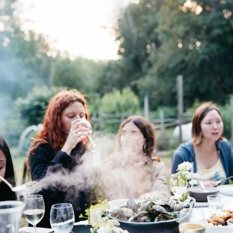A group of women sitting at an outdoor dinner table during sunset, with one woman drinking from a glass and steam rising from a dish of clams on the table.