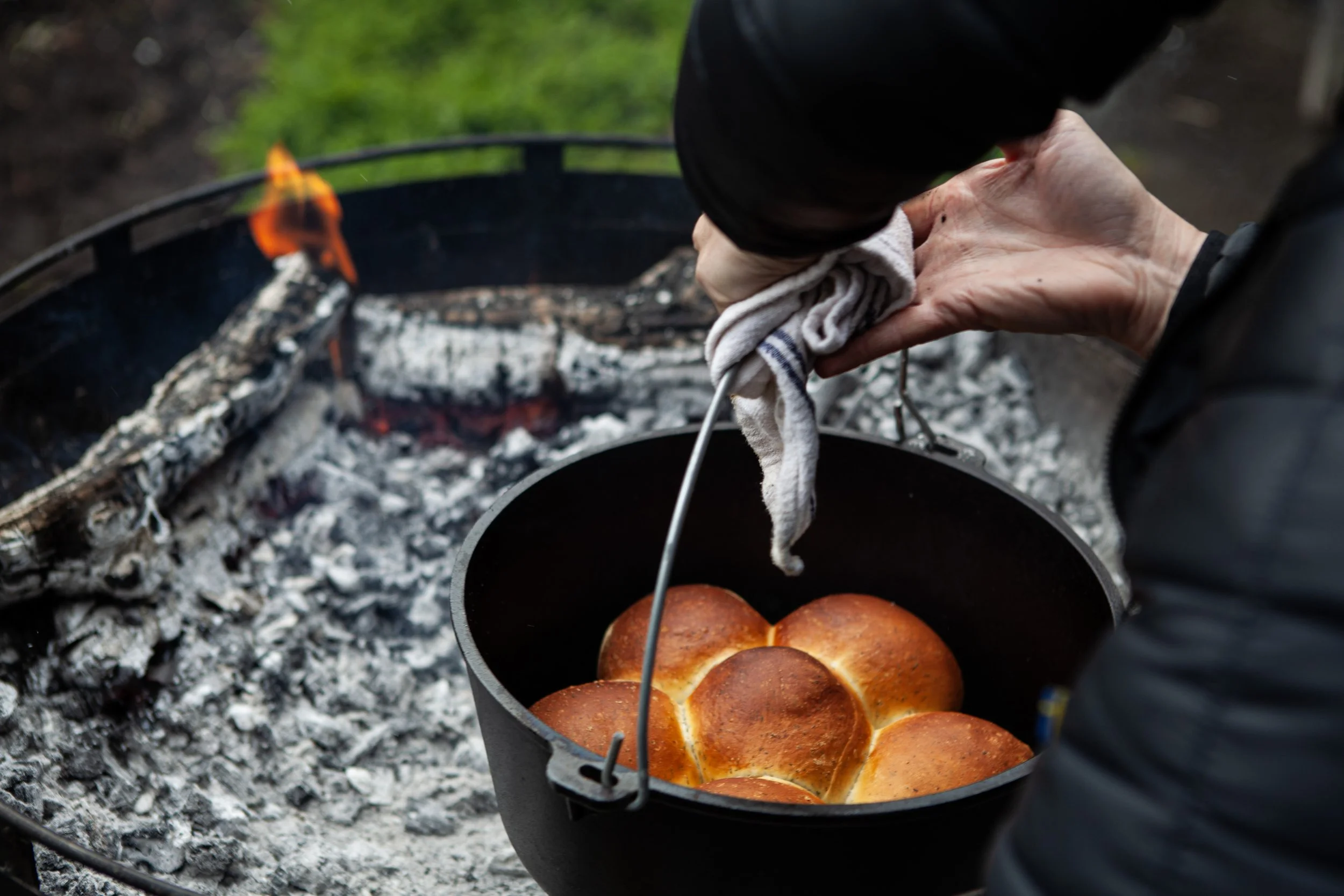 Person removing a hot dish of bread rolls from a charcoal grill with tongs and a cloth outdoors.