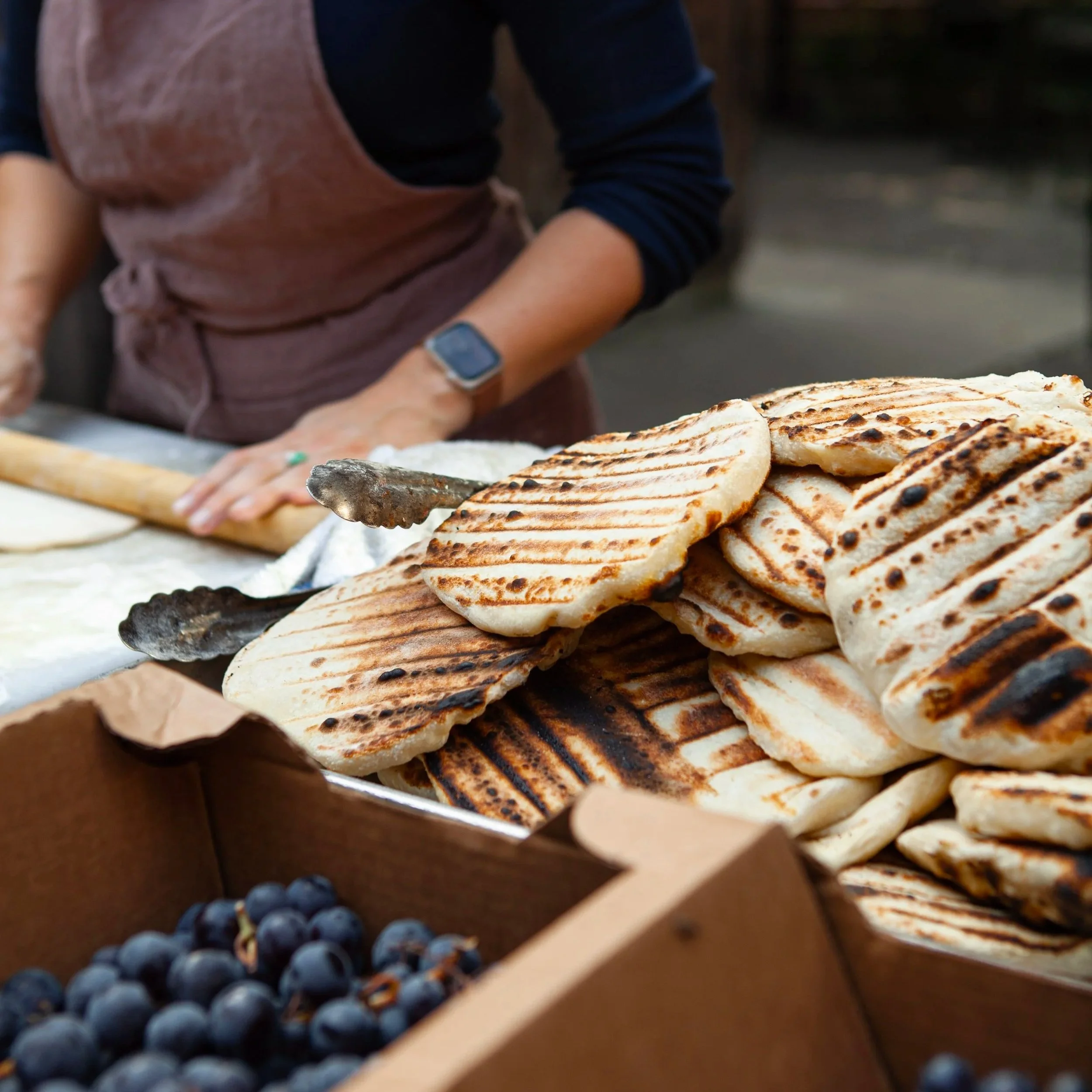 A person preparing grilled flatbread with tongs at an outdoor market, with a box of grapes in the foreground.