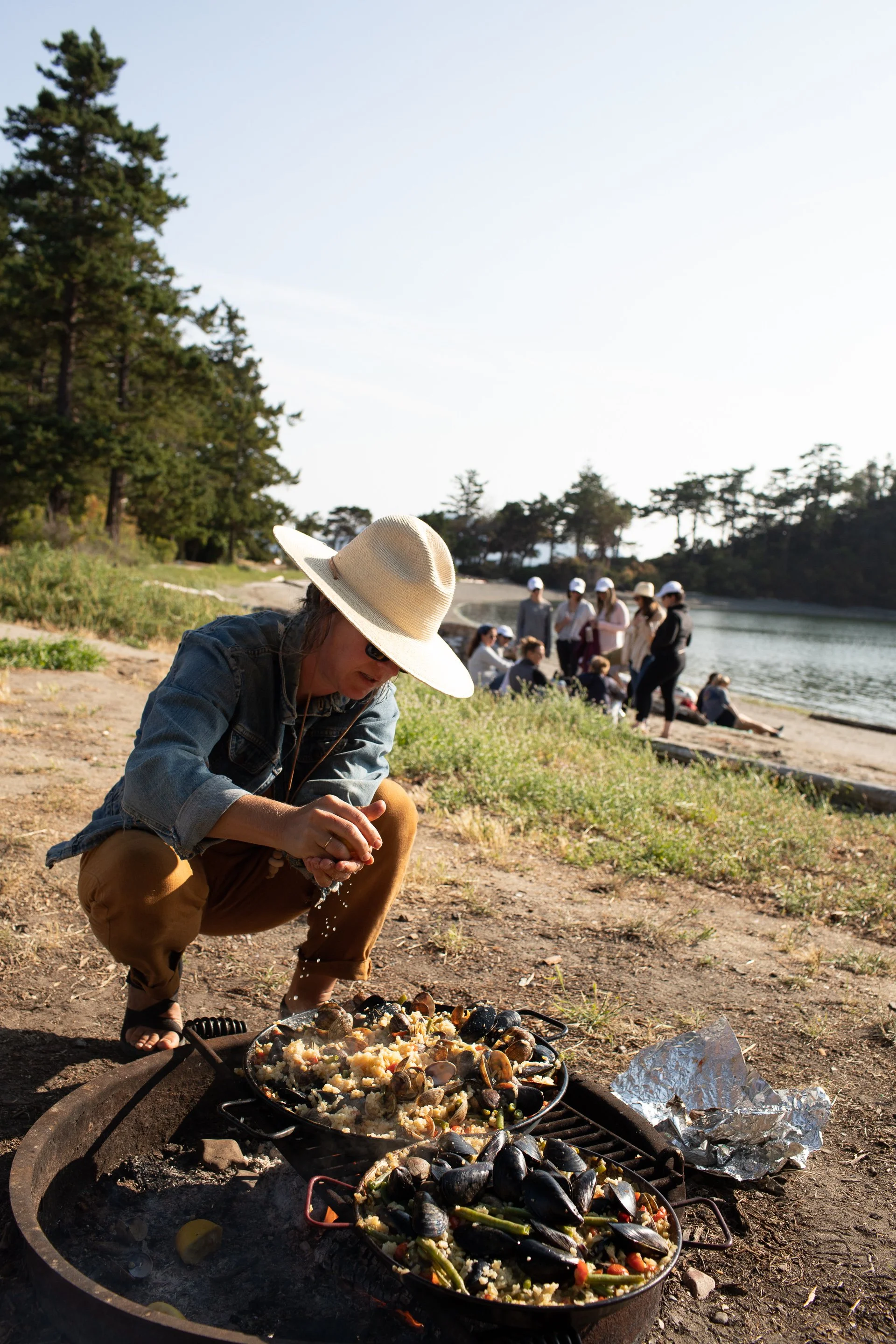 Person wearing a large hat and sunglasses cooking seafood and rice over an open fire on a beach, with a group of people gathered near the water in the background.