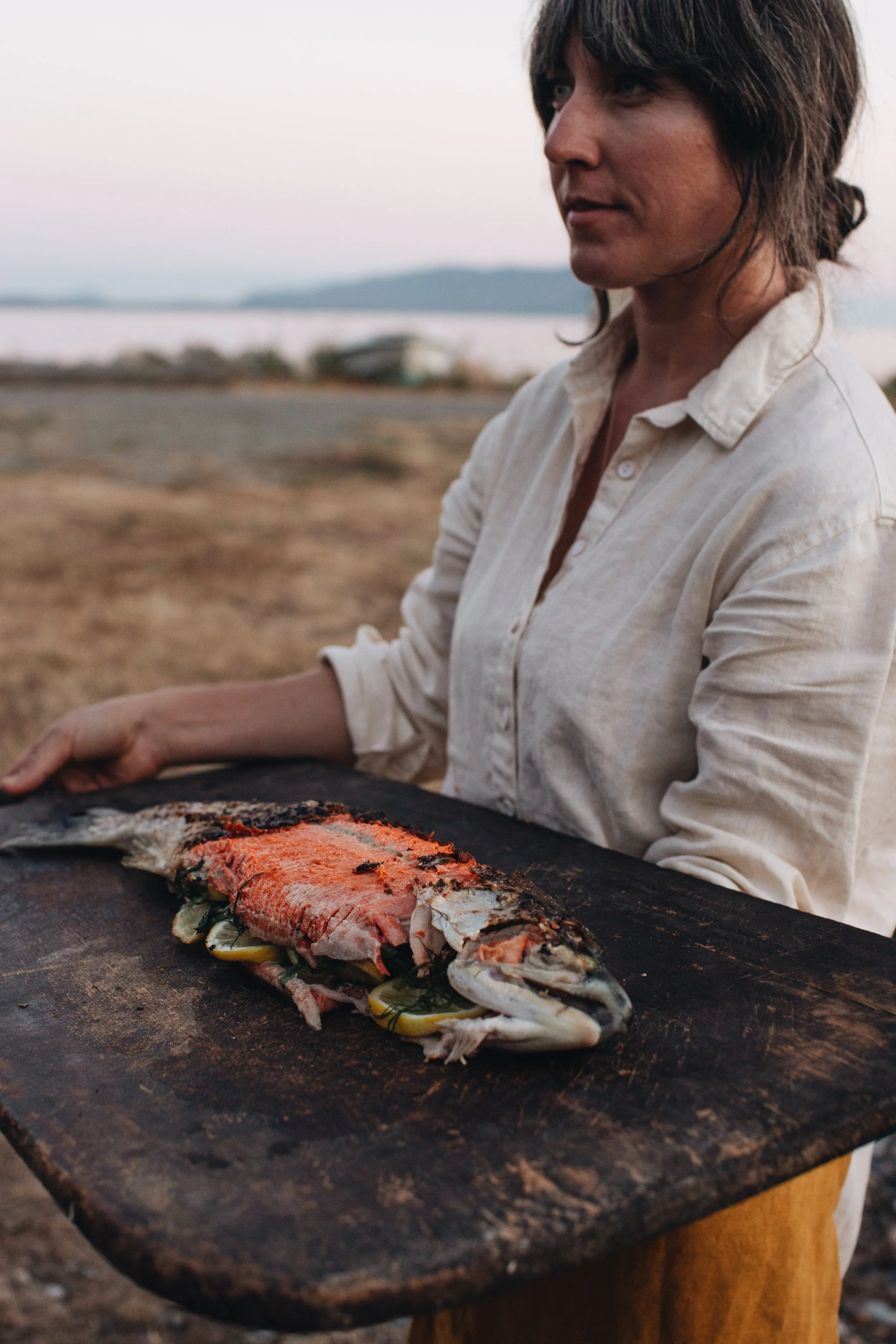 A woman holding a wooden board with a whole cooked fish garnished with vegetables outdoors near a body of water at sunset.