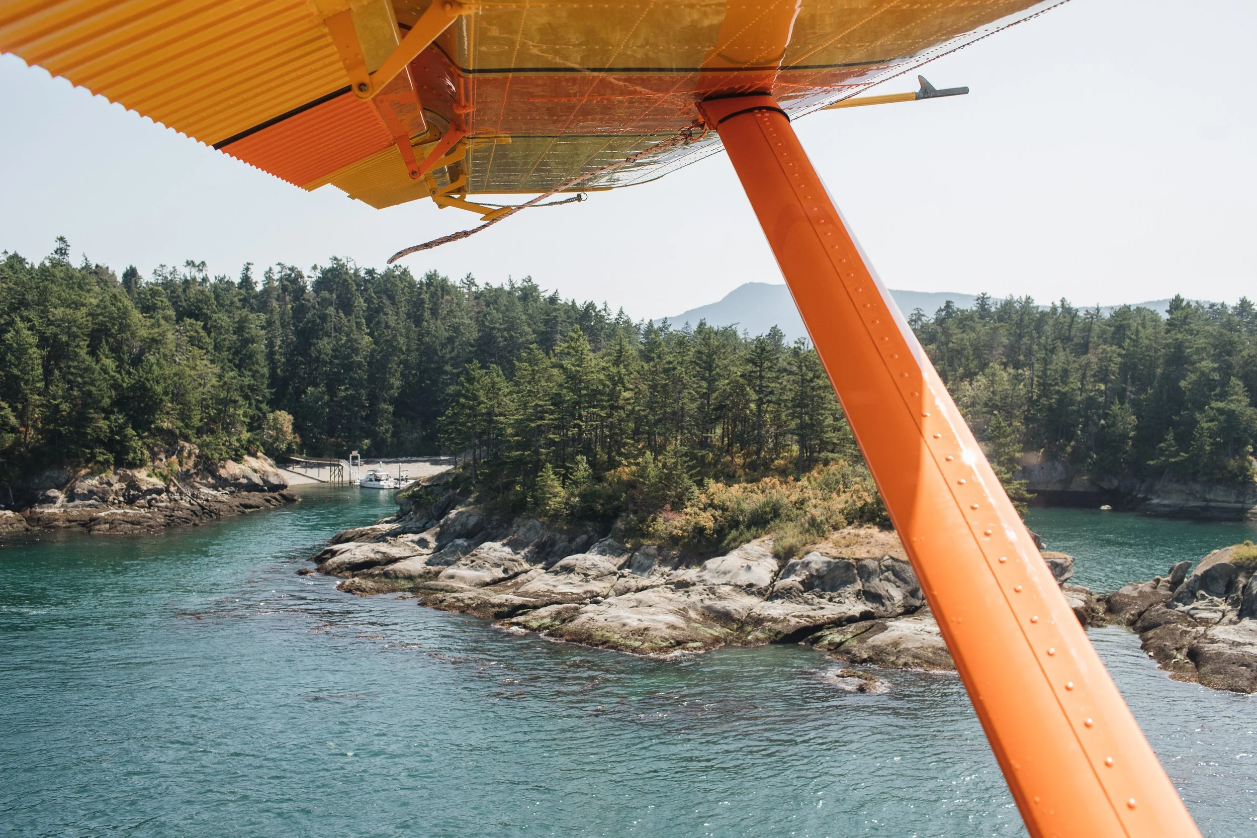 View from a small orange float plane flying over a rocky river surrounded by trees and forest with mountains in the background.