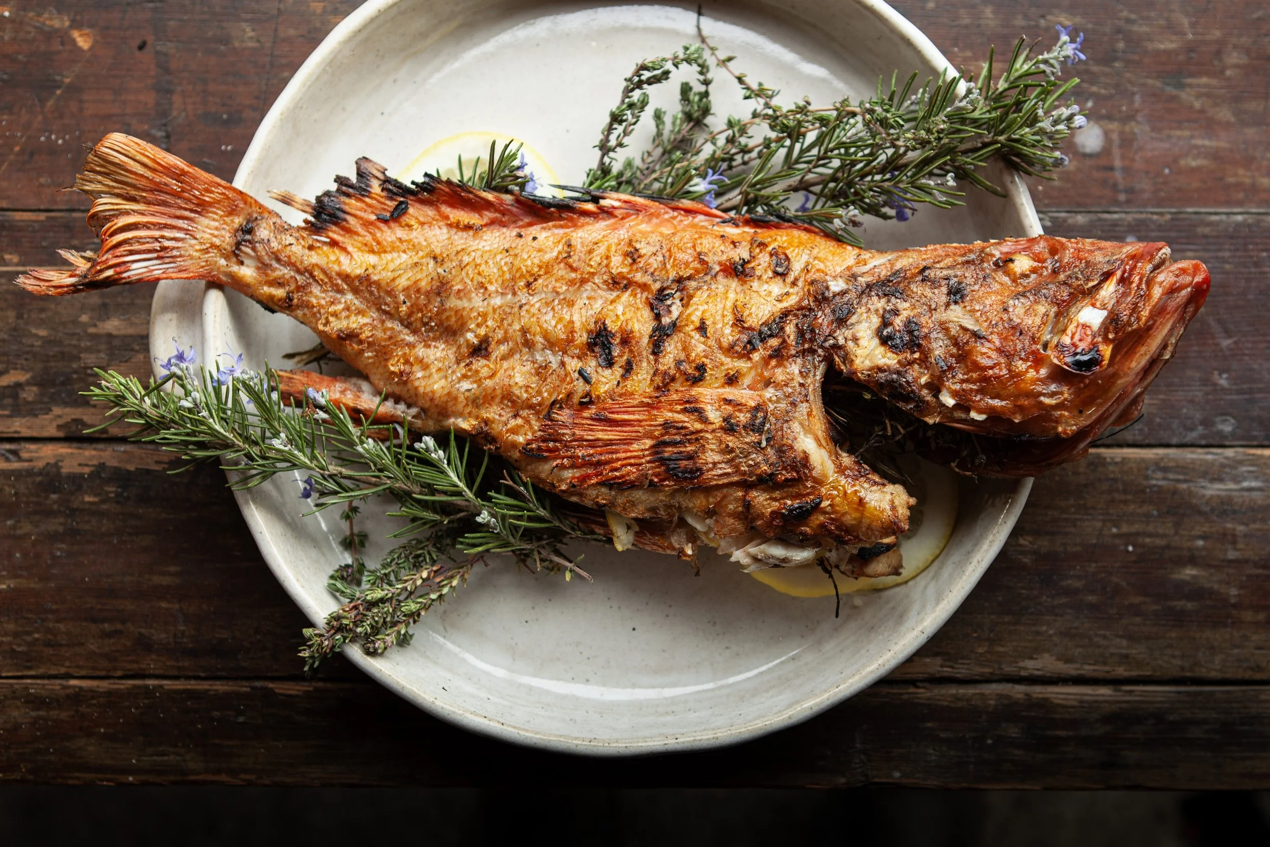 A grilled whole fish on a white plate garnished with rosemary sprigs and lemon slices, placed on a dark wooden table.