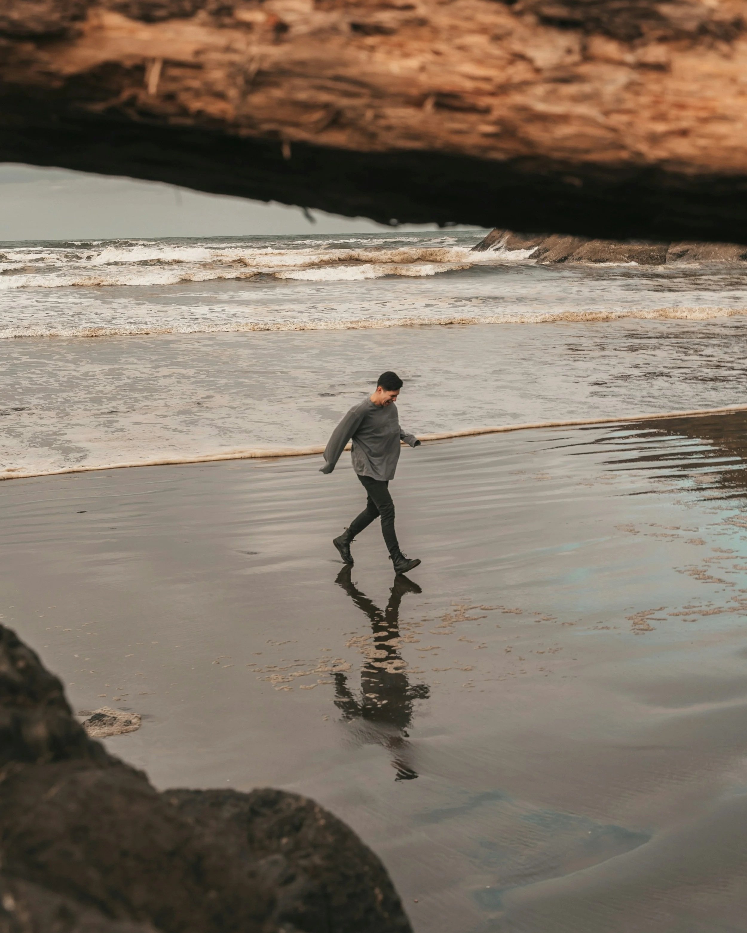 Person walking on a wet beach, seen through rocks or a cave opening, with ocean waves in the background.