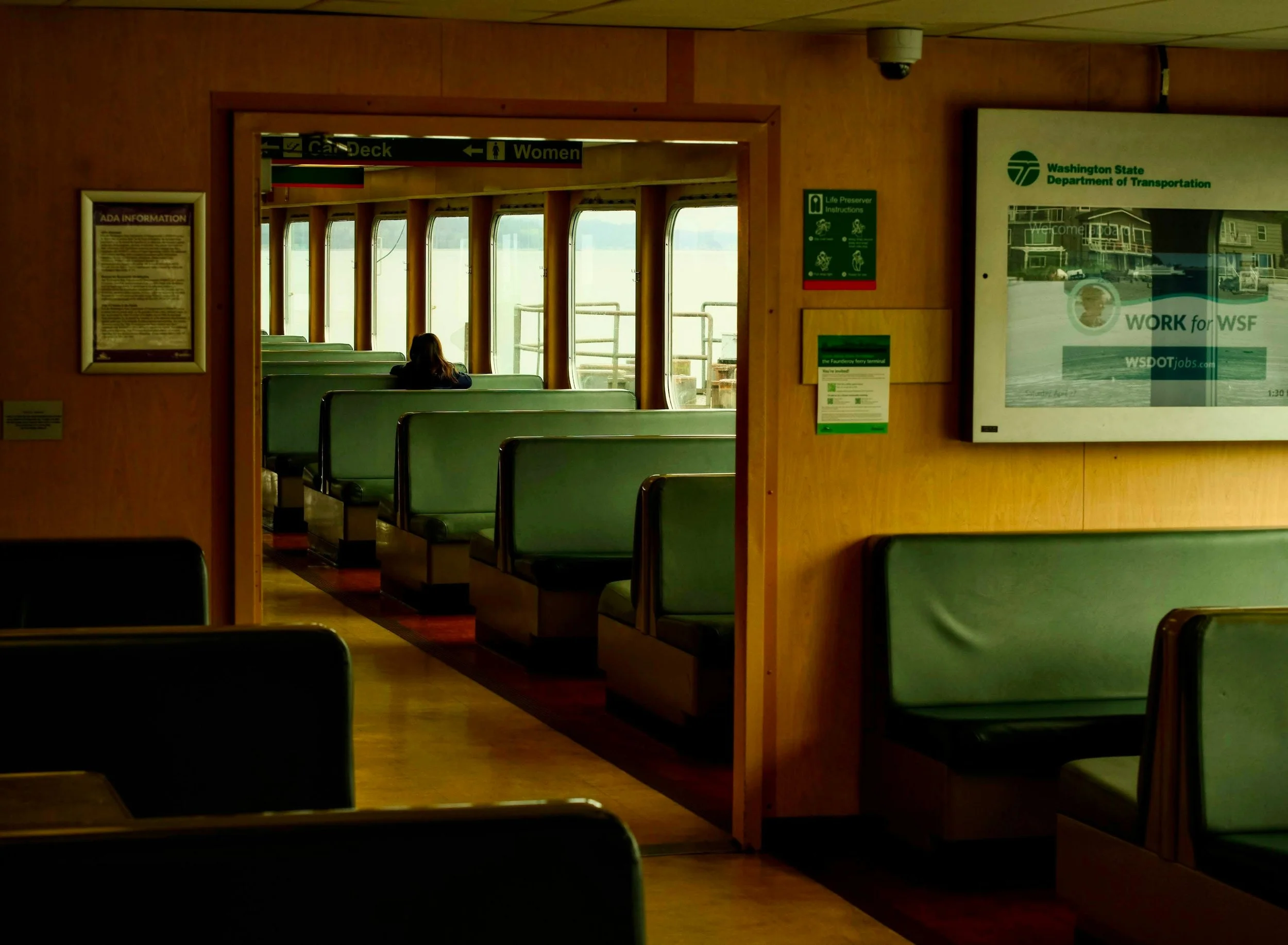 Interior view of a ferry passenger area with green seats, wooden walls, and windows showing water outside; a woman is sitting alone, and signs indicating deck, women, and other information are visible.