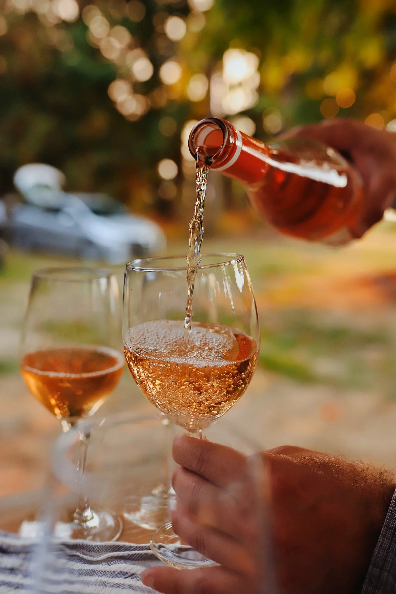 Person pouring rosé wine into a glass with a second glass of rosé wine nearby, set outdoors with a blurred background of trees and a car.