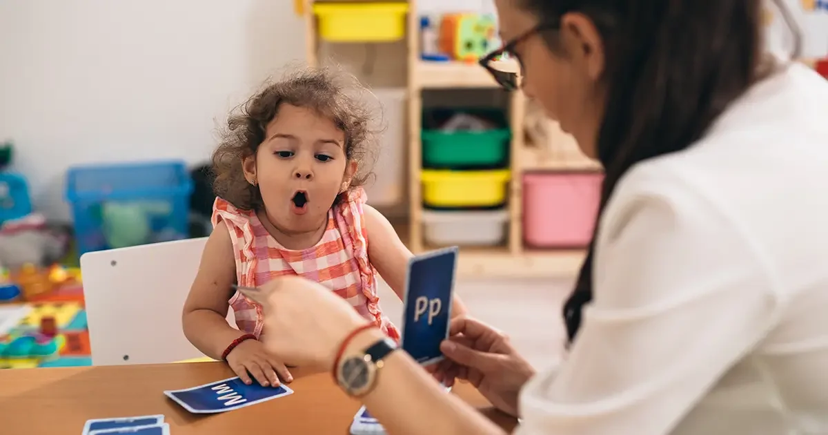 A young girl sitting at a table with a surprised expression as she learns phonics, with a teacher holding a flashcard showing the letter P.