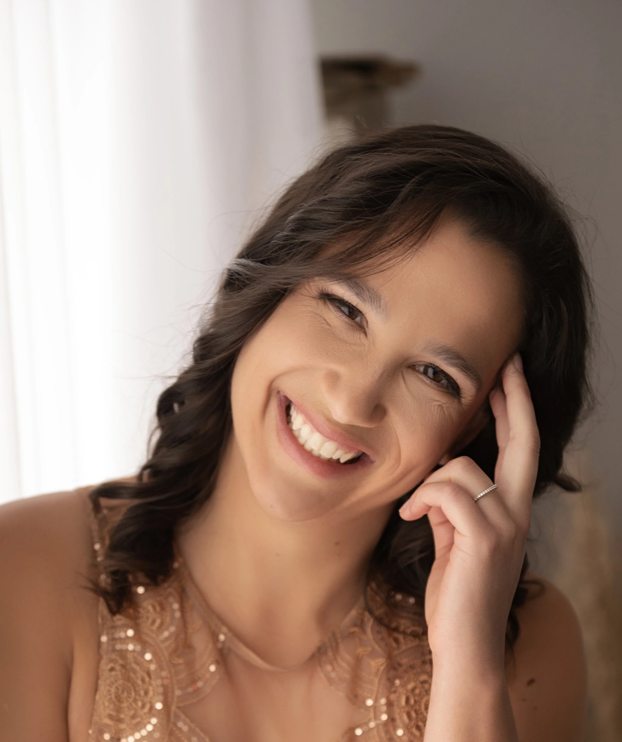 A young woman with short brown hair smiling and leaning her head on her hand, wearing a peach-colored dress with intricate lace detailing.