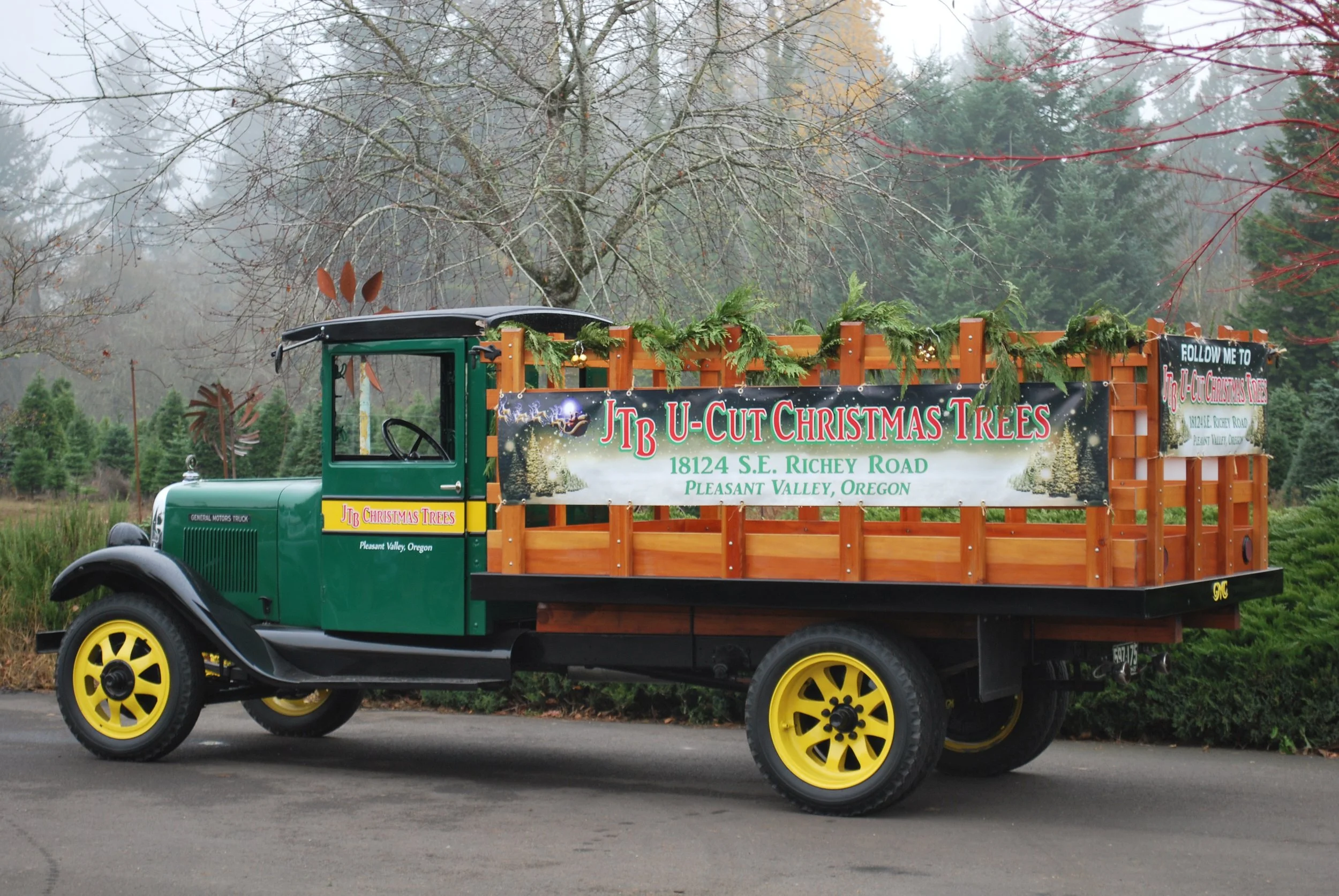 Vintage green truck decorated with Christmas greenery and ornaments, promoting JTB U-Cut Christmas Trees at 18124 S.E. Richey Road, Pleasant Valley, Oregon, parked outdoors with leafless trees and evergreen trees in the background.