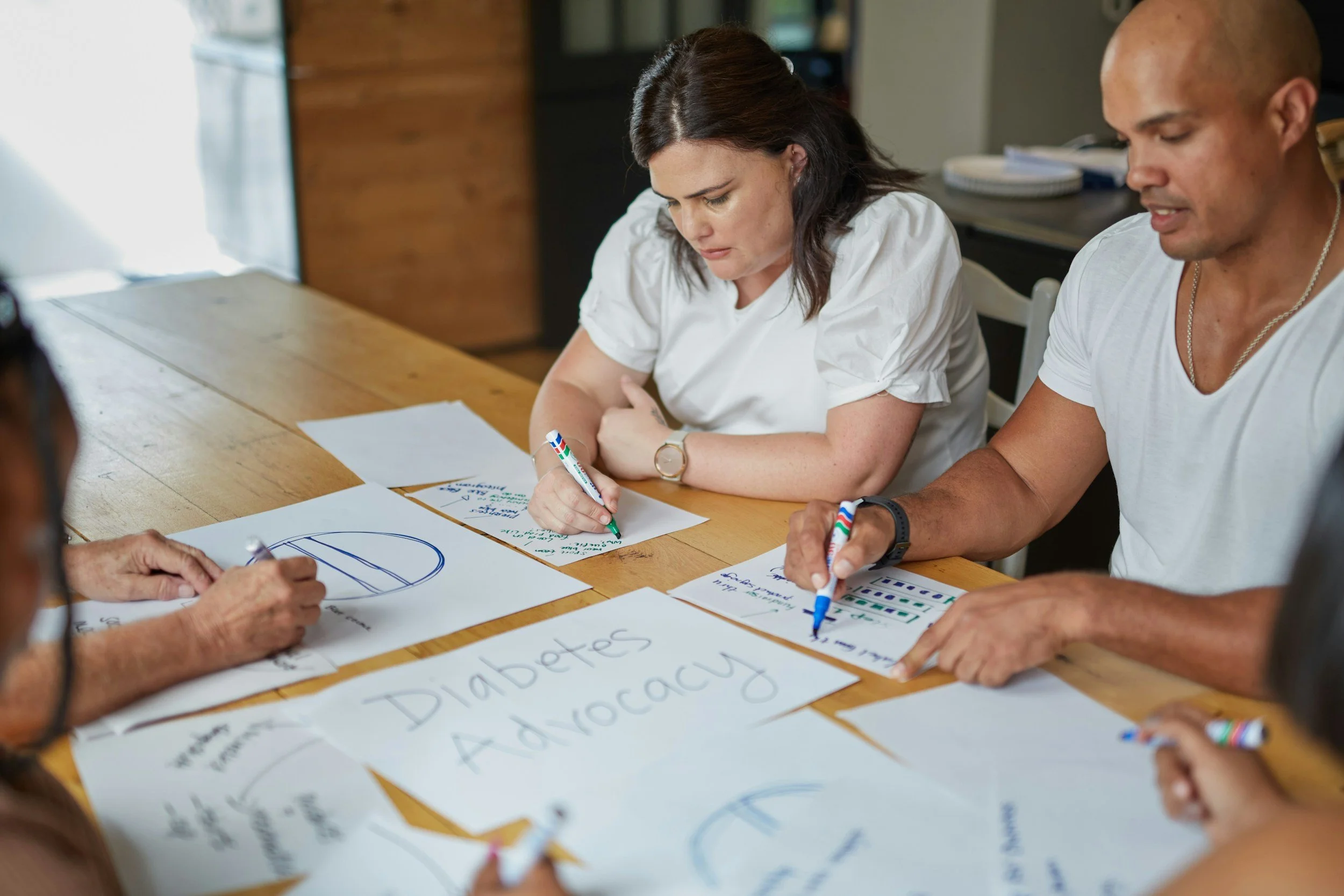People sitting around a wooden table working on papers related to diabetes advocacy, with some papers showing charts and a sign that says "Diabetes Advocacy".