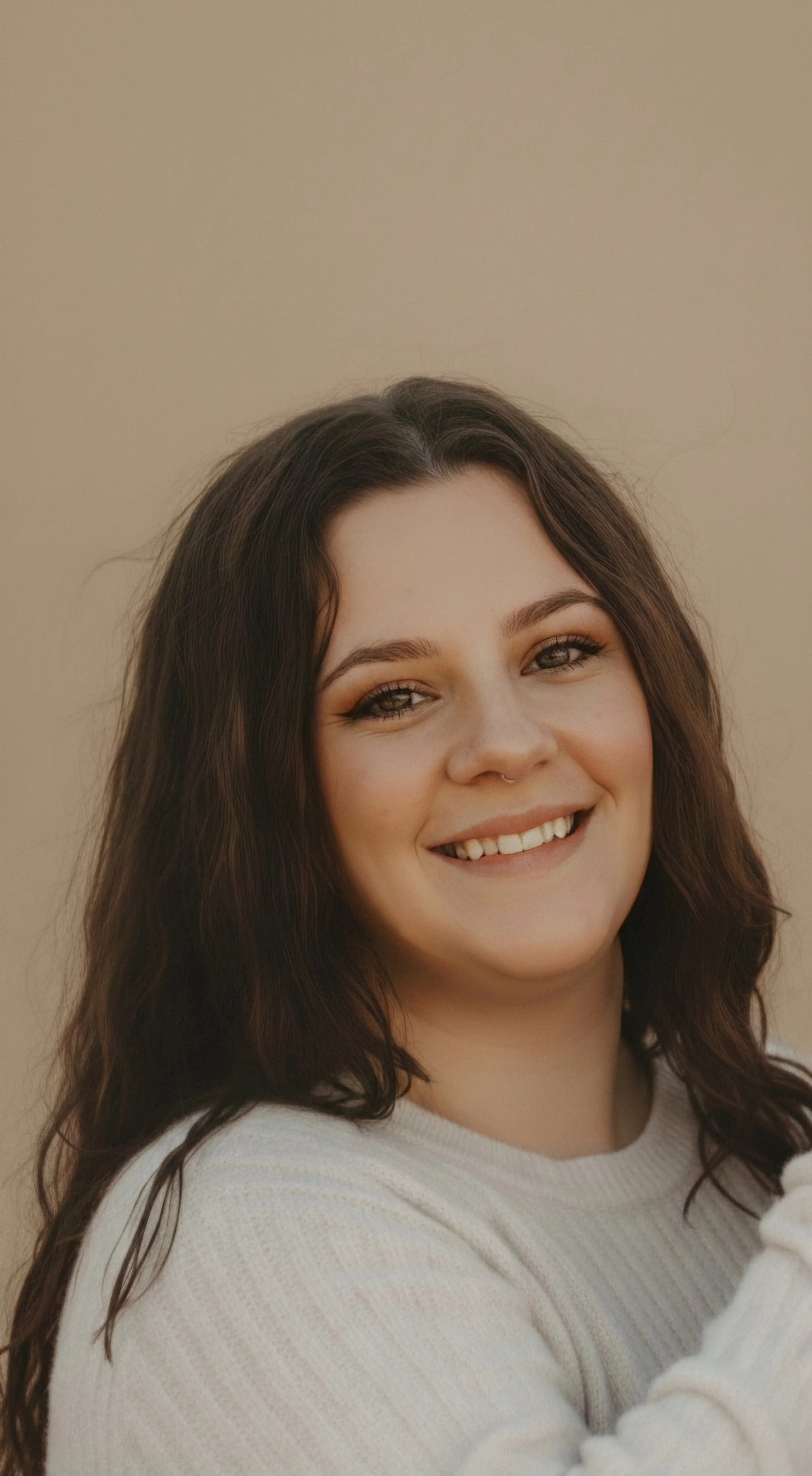 Close-up portrait of a young woman with dark wavy hair, smiling softly, wearing a white sweater, standing against a plain beige background.