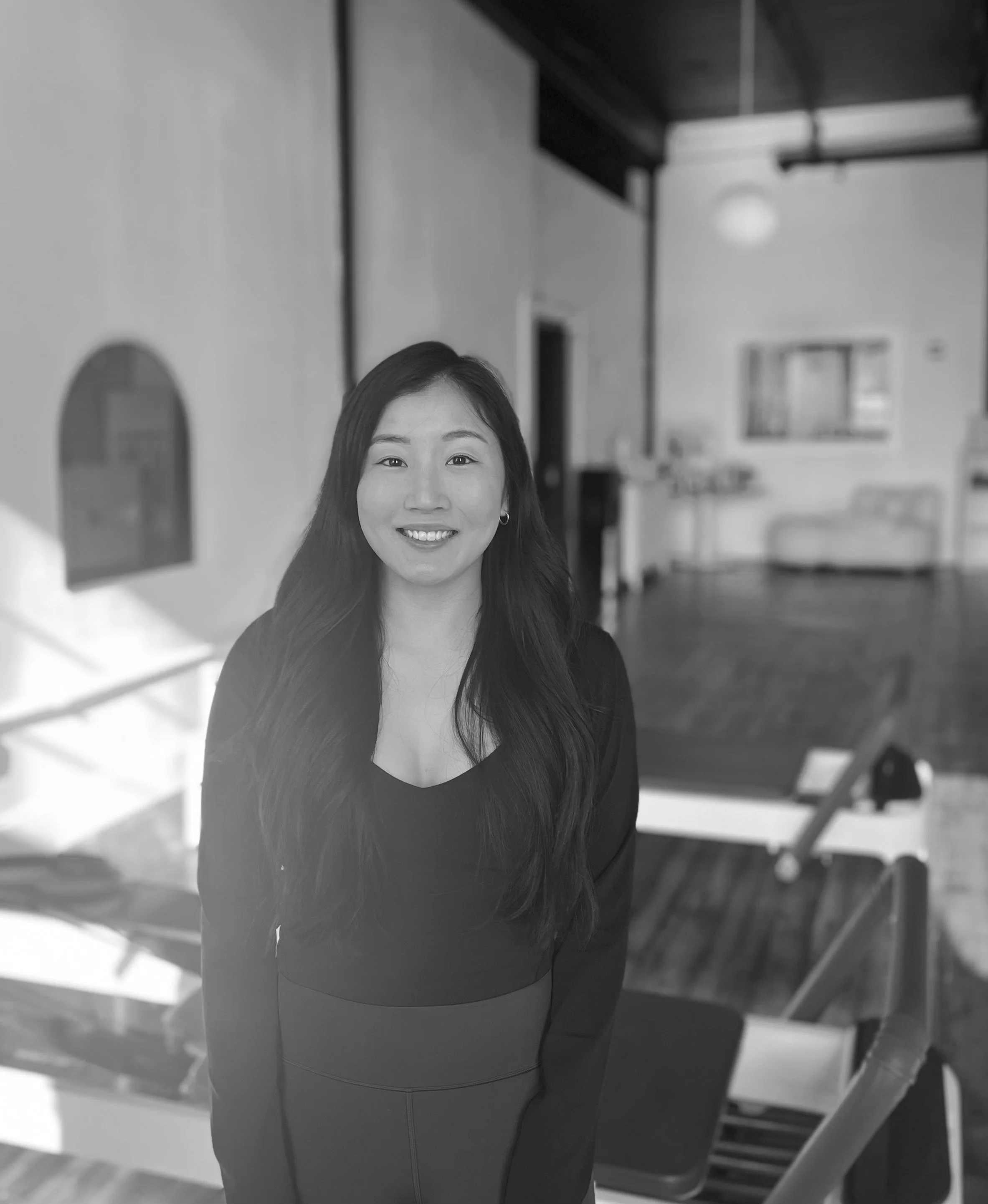 Grayscale photo of a smiling woman with long dark hair, wearing a black top and a dark skirt, standing in a spacious room with wooden floors and large windows in the background.