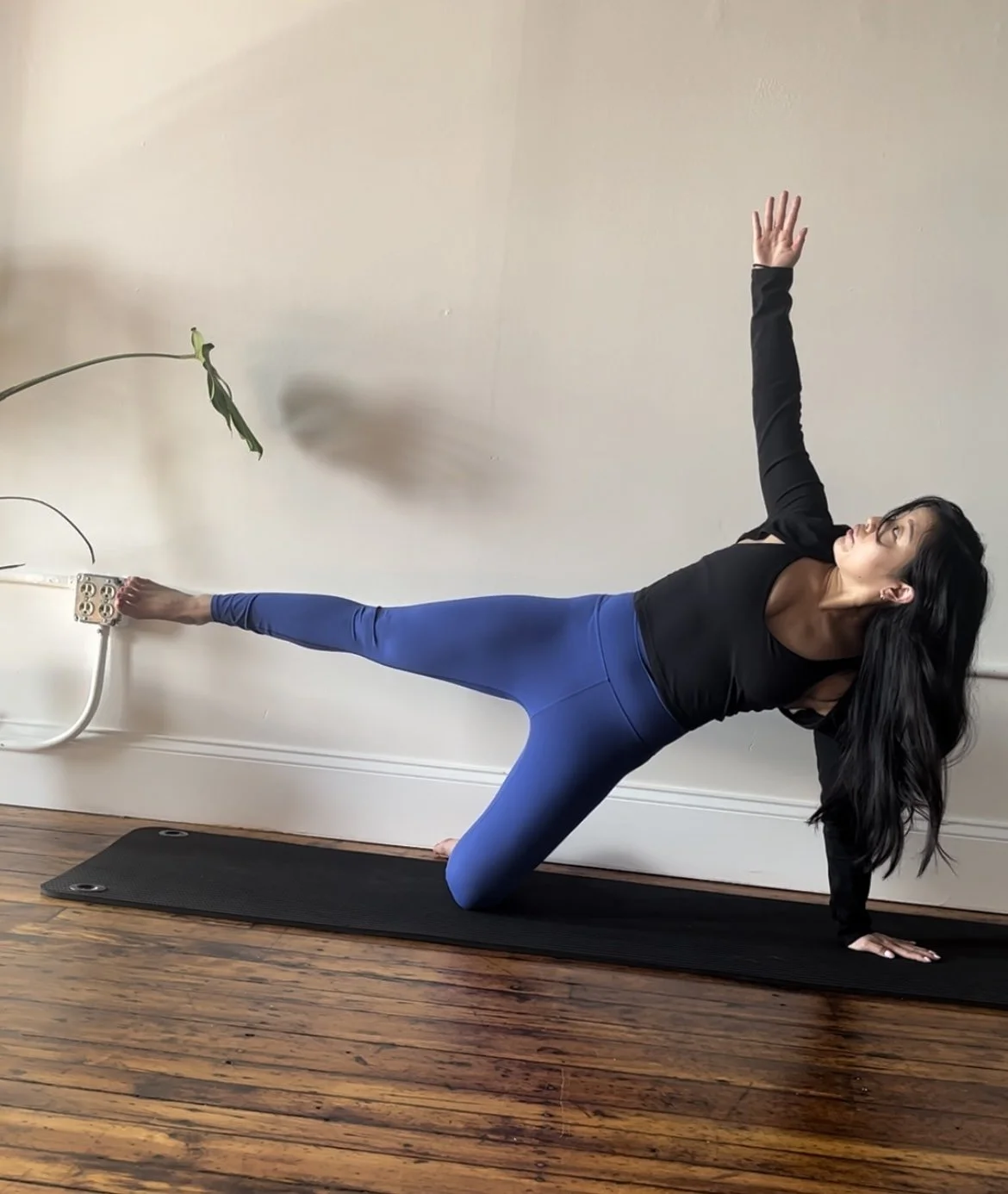 Woman performing a side plank pilates pose on a black mat in a room with hardwood floors, wall socket, and an indoor plant.