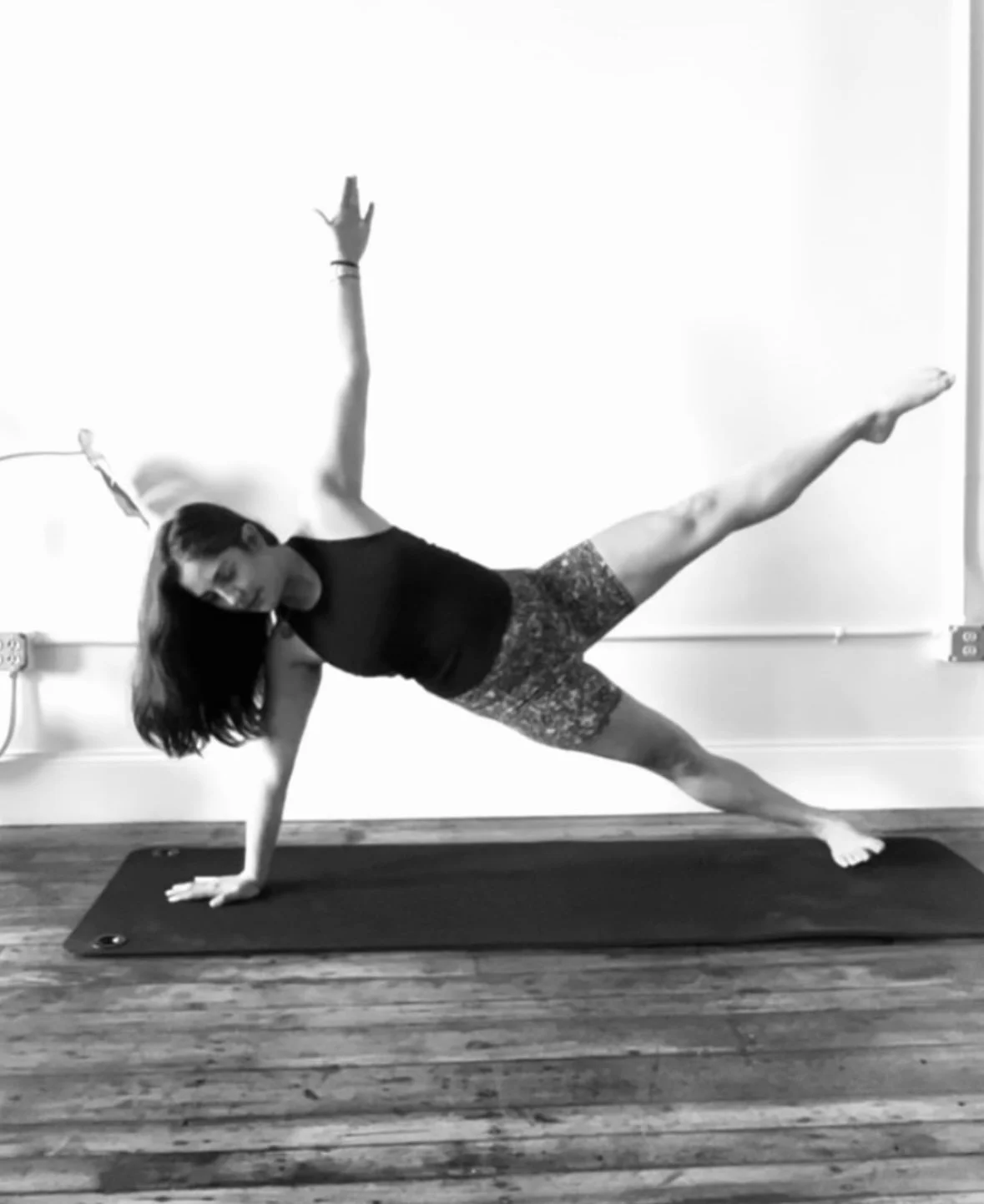 Woman performing a side plank pilates pose on a black mat in a room with wooden floor, wearing a black top and patterned shorts.