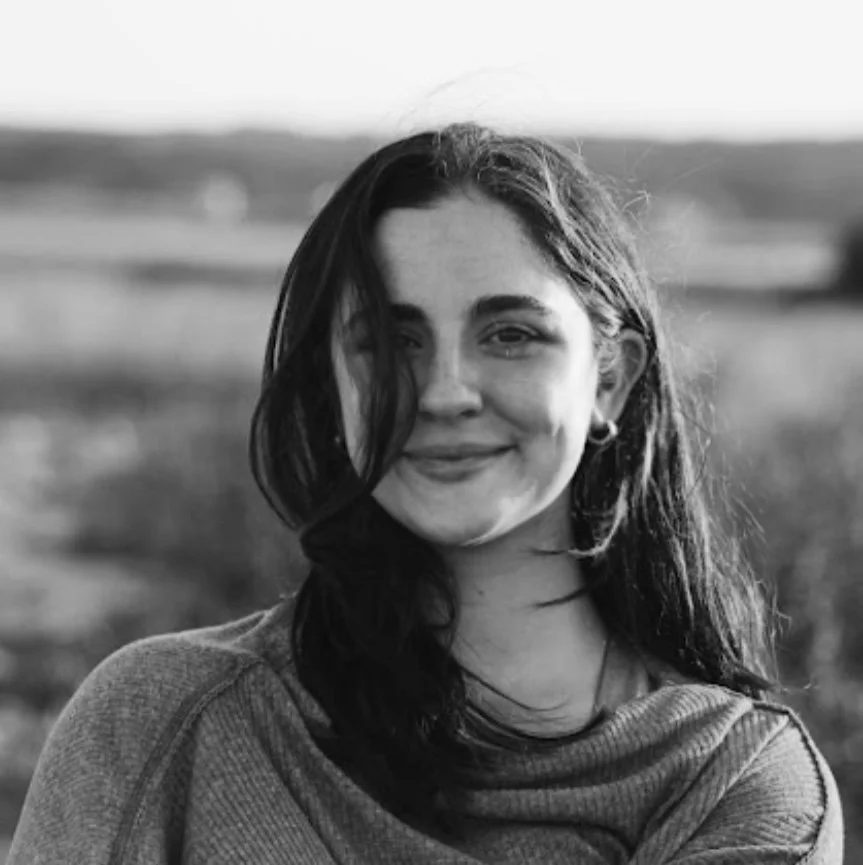 A young woman outdoors with a smile, in black and white, with a blurred landscape background.