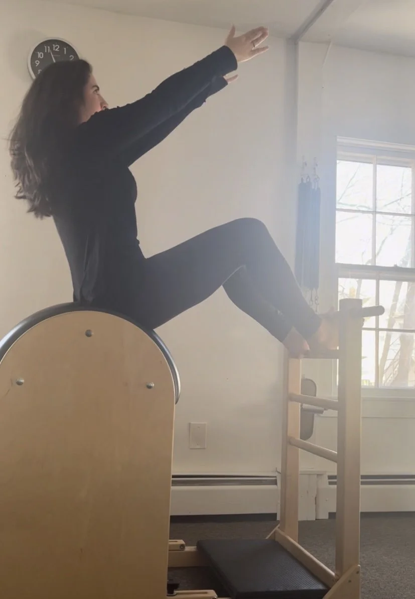 A woman in black exercise clothes sitting on a Pilates ladder barrel with her legs extended forward and raised, reaching towards the ceiling with her arms, inside a bright room with a window and a wall clock.