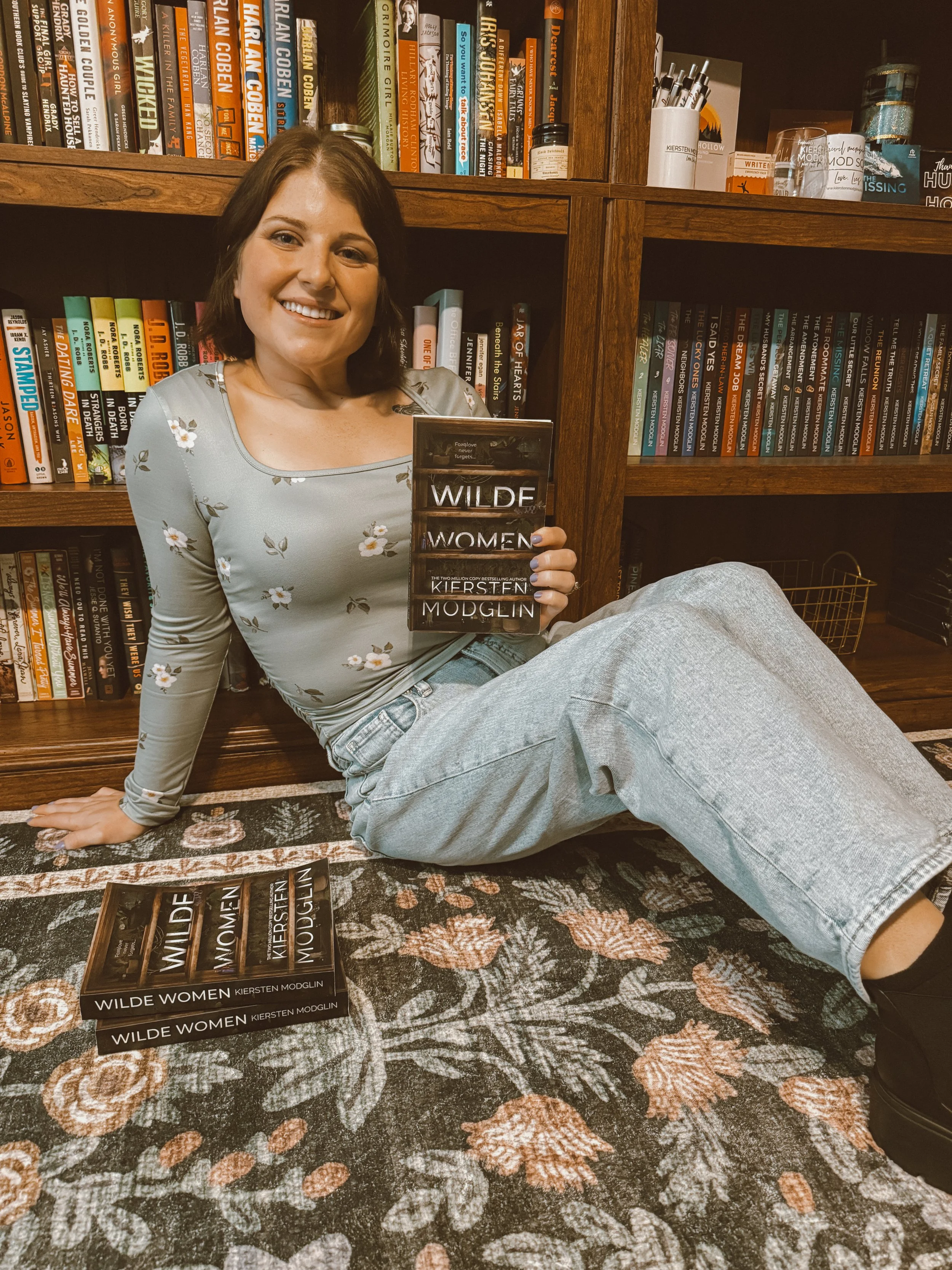 A woman sitting on a floral rug in front of a bookshelf, holding a book titled 'Wilde Women' by Kiersten Modglin. The bookshelf contains various books, and several copies of the same book lie on the rug in front. The woman is smiling and wearing a light gray floral long-sleeve top and light blue jeans.
