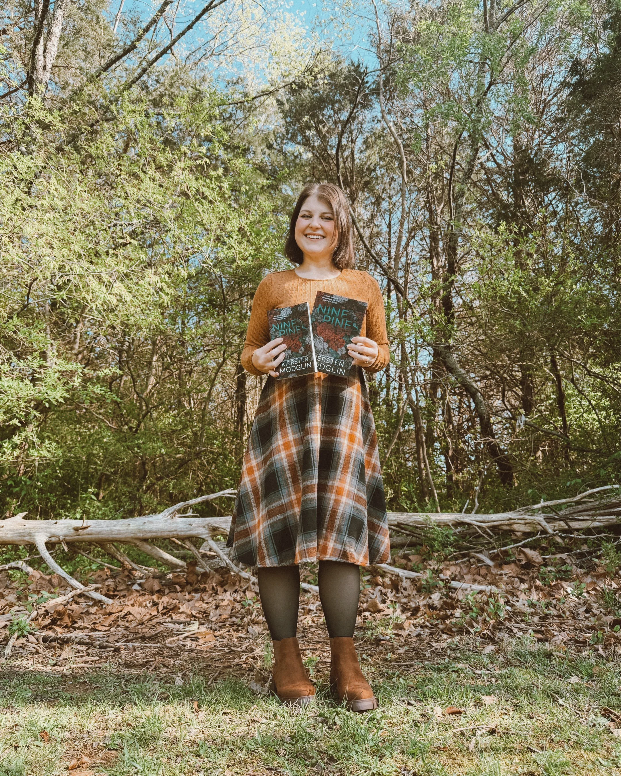 A woman holding two copies of a book titled 'Nine Pines' by Kiersten Modglin outdoors in a wooded area, smiling, with trees and a fallen log in the background.