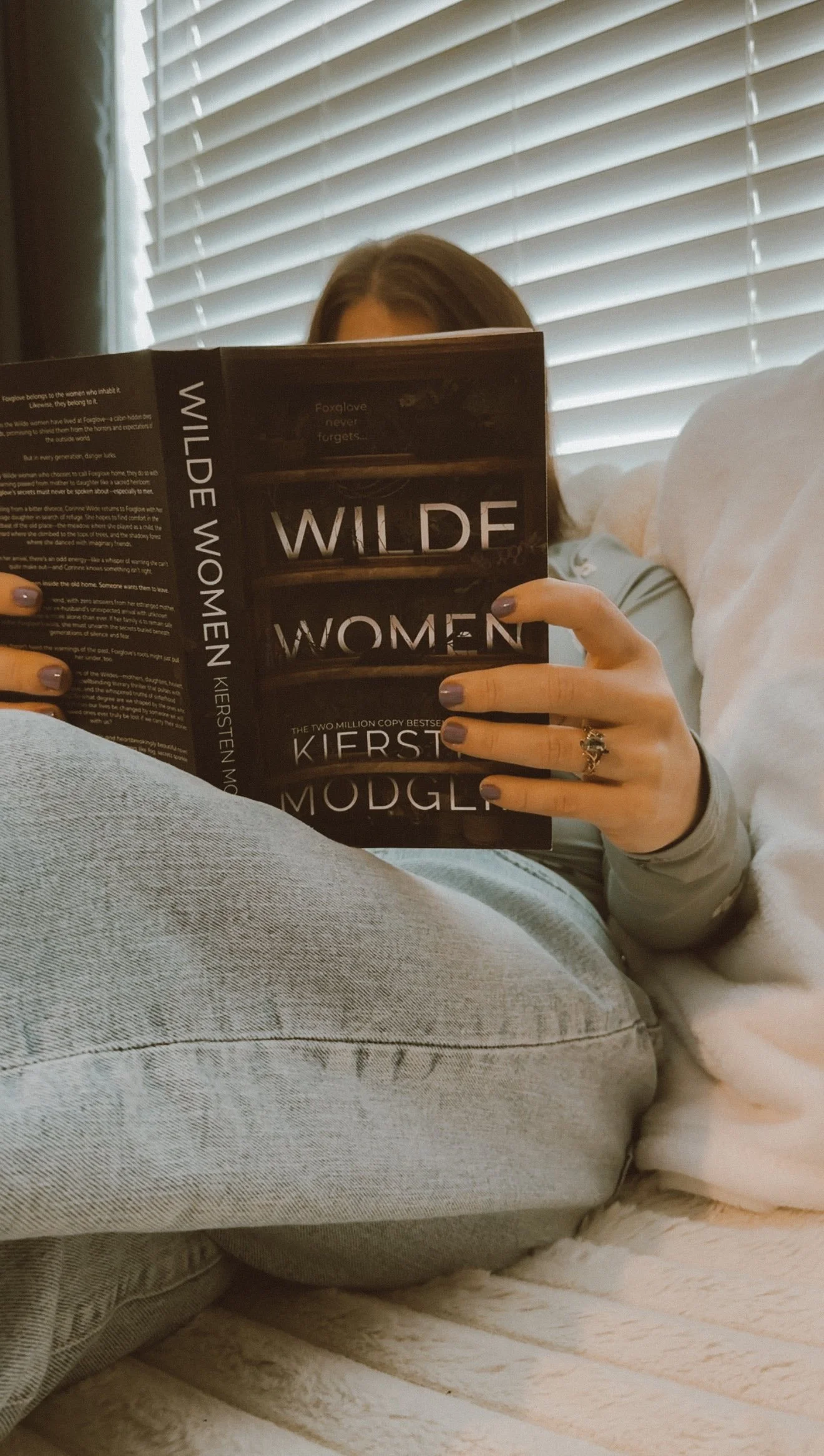 A woman sitting on the floor and reading a book titled 'Wilde Women' by Kiersten Modglin, with her face hidden behind the book, on a cozy indoor setting with closed blinds in the background.