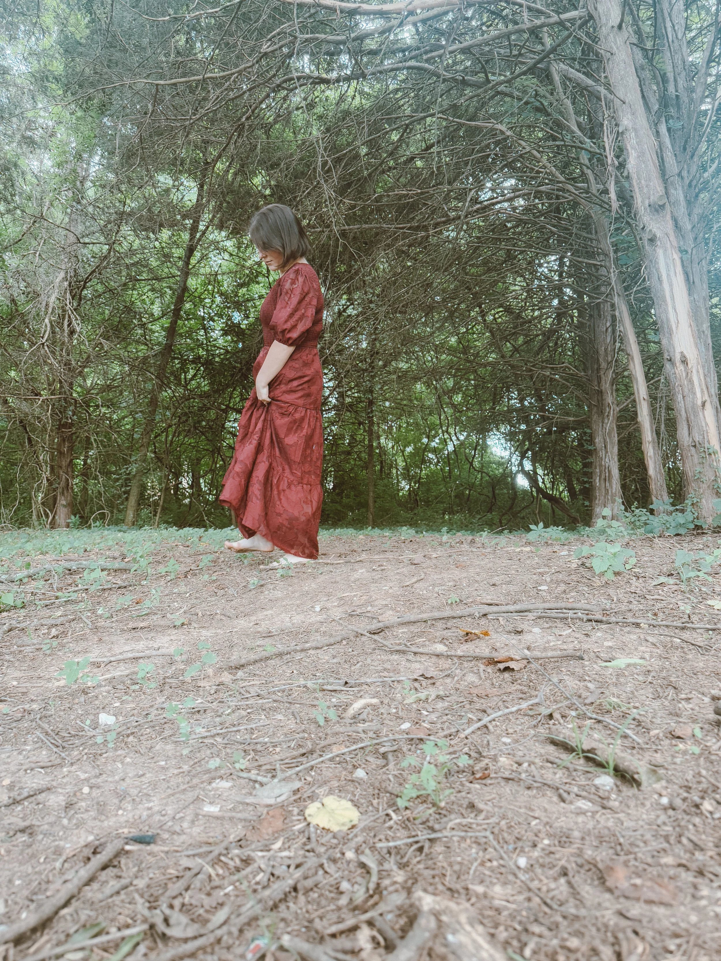 A woman in a long red dress standing barefoot on dirt ground in a dense forest, looking down.