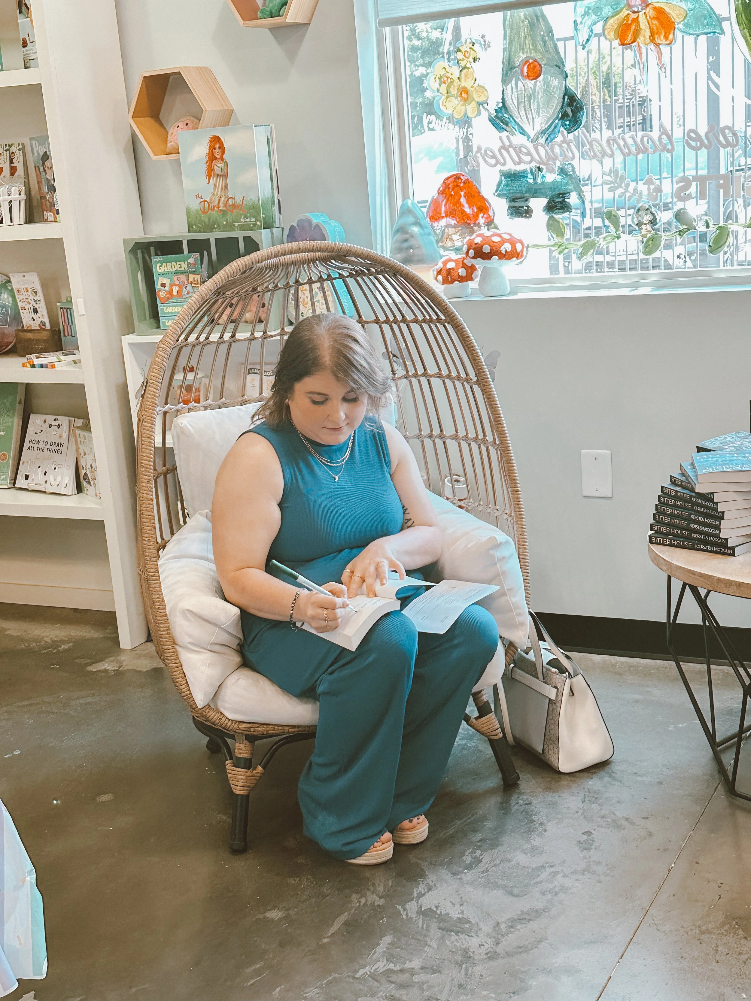 A woman with brown hair, wearing a blue sleeveless top and teal pants, sits on a wicker chair with a white cushion, writing in a notebook. The setting is a cozy bookstore or cafe with a shelf of books, decorative items, and colorful mushroom decorations on the window sill.