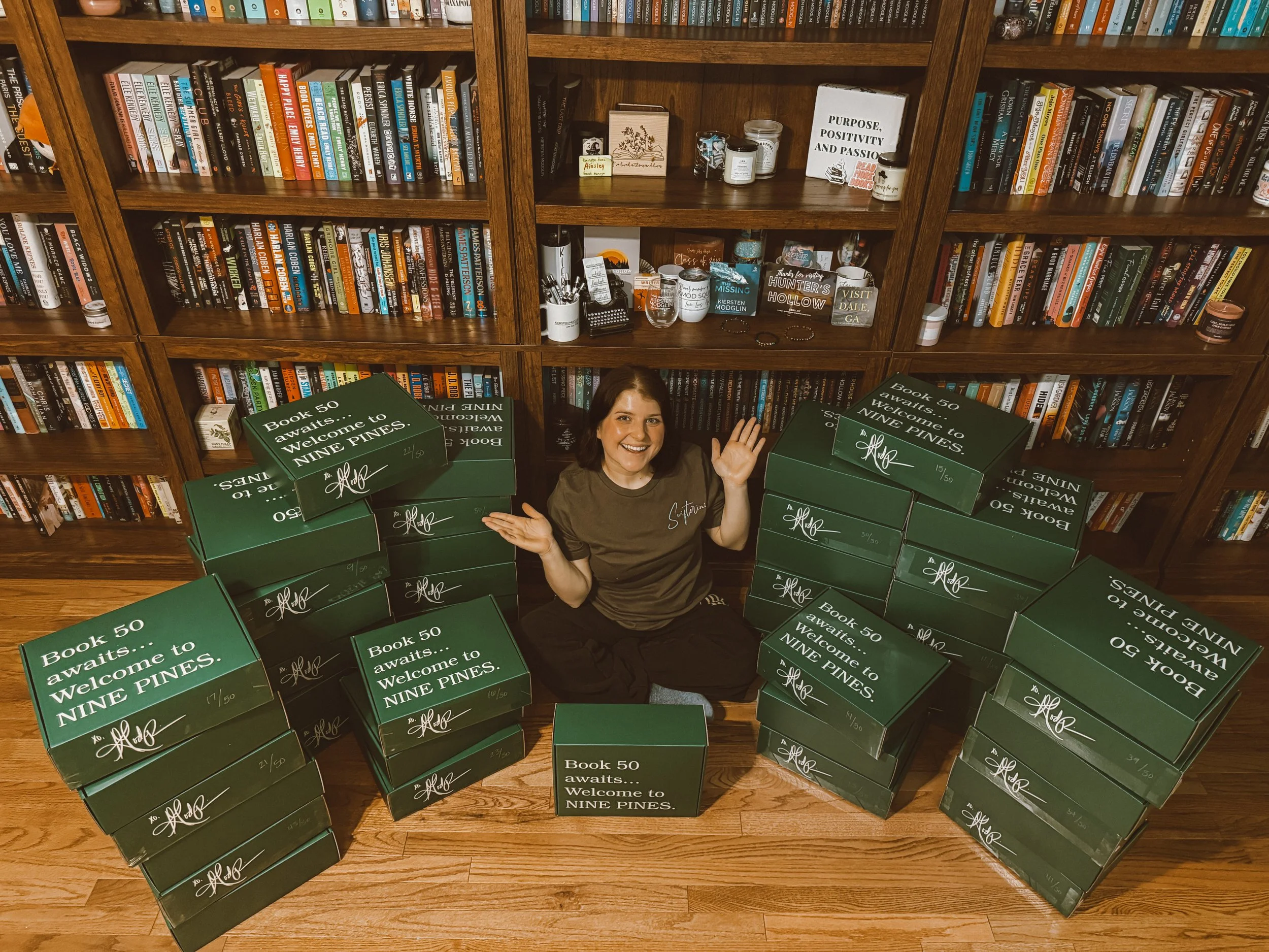 Kiersten Modglin sitting on the floor in front of bookshelves, surrounded by stacks of green boxes labeled 'Book 50 awaits... Welcome to NINE PINES,' smiling and waving at the camera.