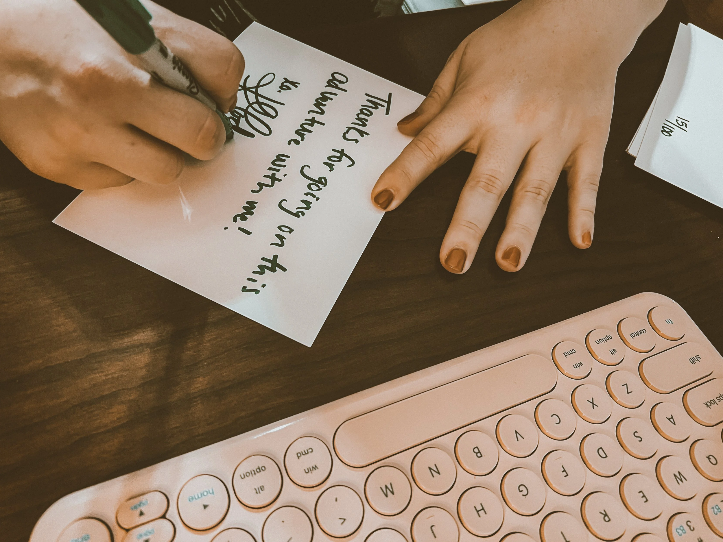 Person writing a message on a white piece of paper with a black marker, with a pink keyboard nearby on a brown wooden desk.