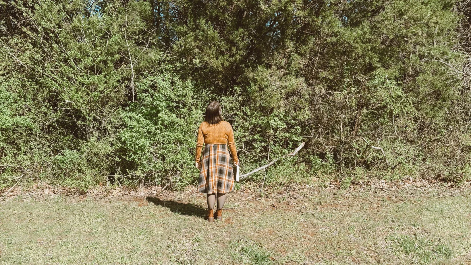 A woman with brown hair wearing a tan sweater, plaid skirt, and brown boots stands facing a green bushy area with her back to the camera, holding an object in her right hand.