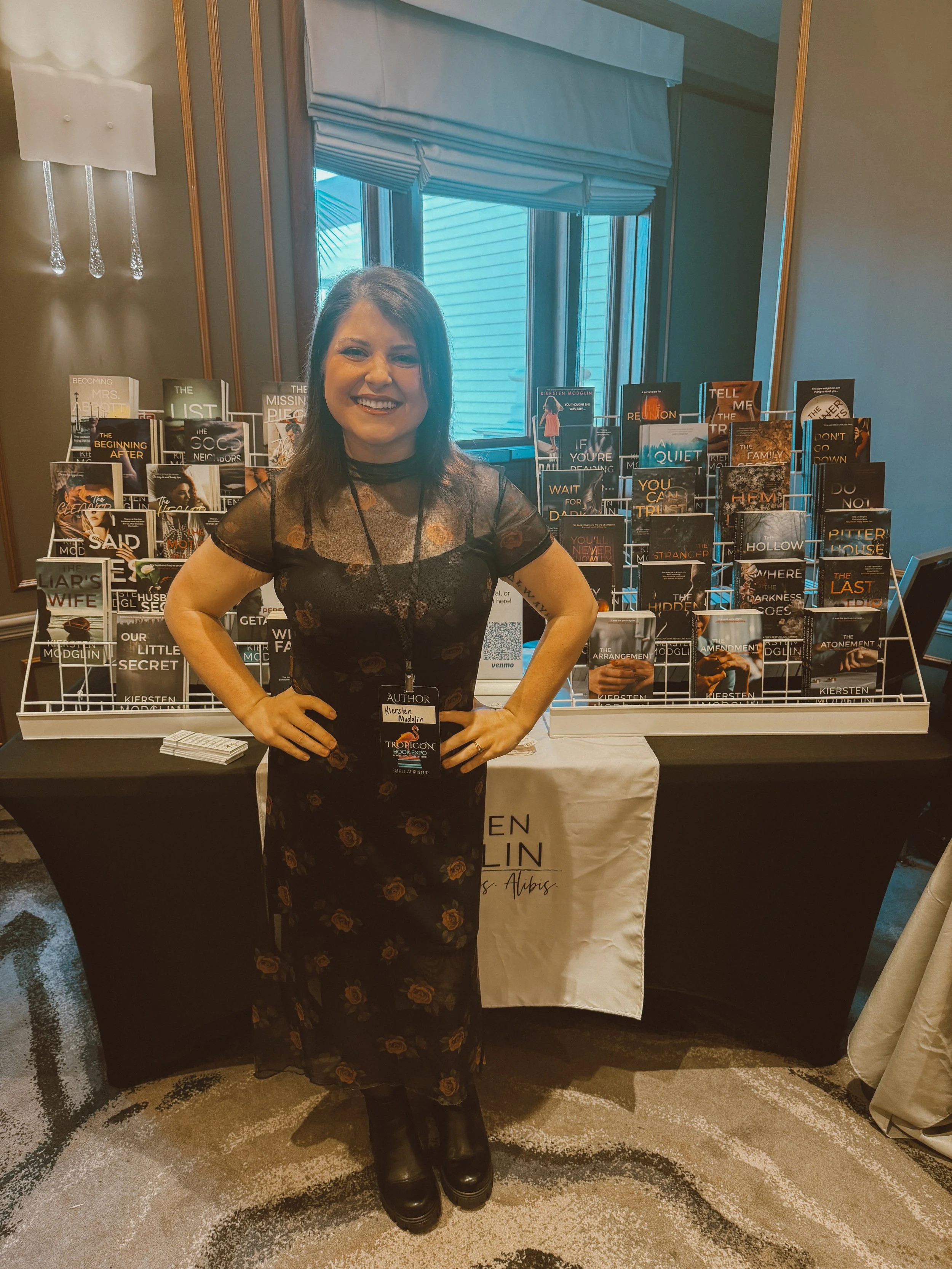 A woman standing in front of a table with numerous books displayed on a rack. She is smiling, wearing a black floral dress with sheer sleeves, black boots, and a conference badge that says 'Author.' The background features a window with blinds and a white banner on the table with text.