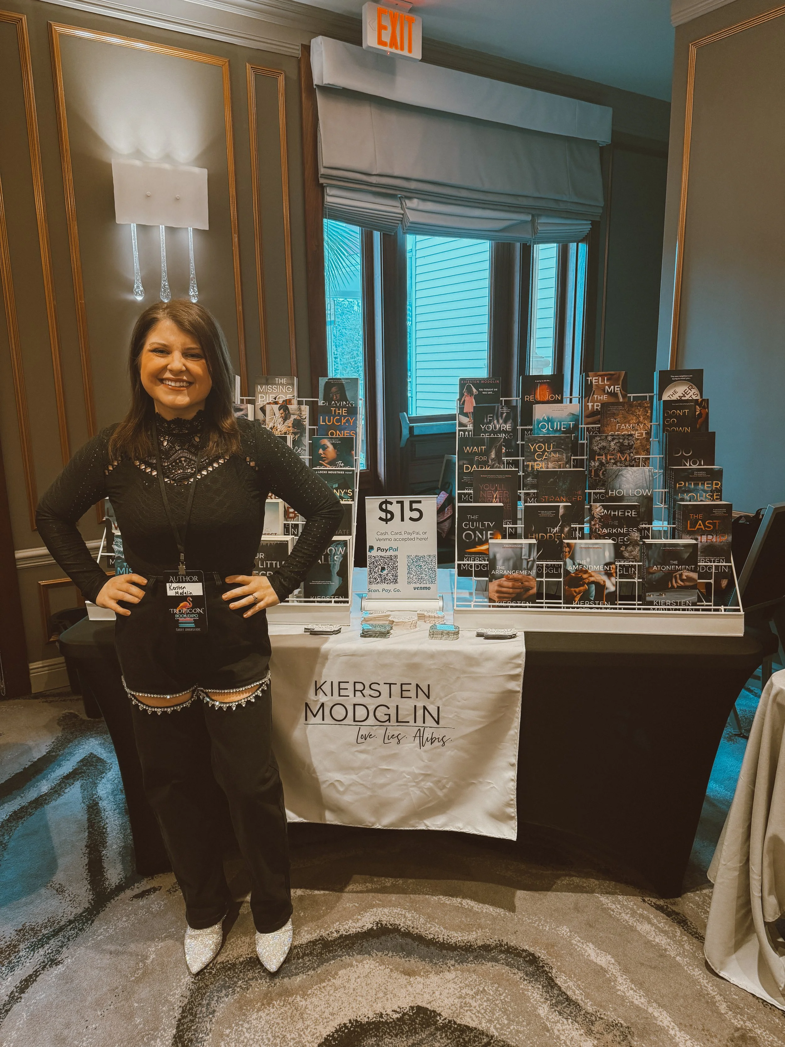 A woman stands in front of a table displaying books at a signing event for Kiersten Modglin, with a sign indicating a price of $15, at an indoor venue with window and exit sign in the background.