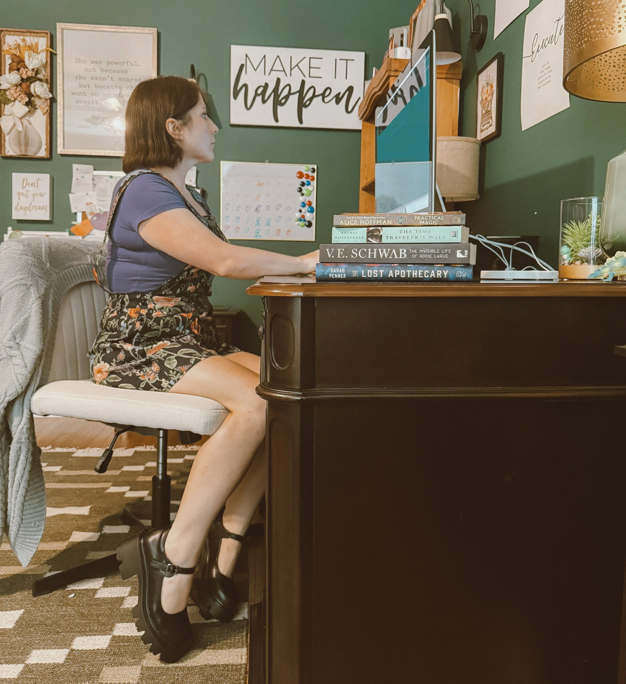 A woman sitting at a desk, working on a computer. She has short brown hair, is wearing a blue top, floral shorts, and black chunky shoes. The desk has a stack of books, a monitor, and some decorative items. The room has green walls decorated with framed quotes and artwork.