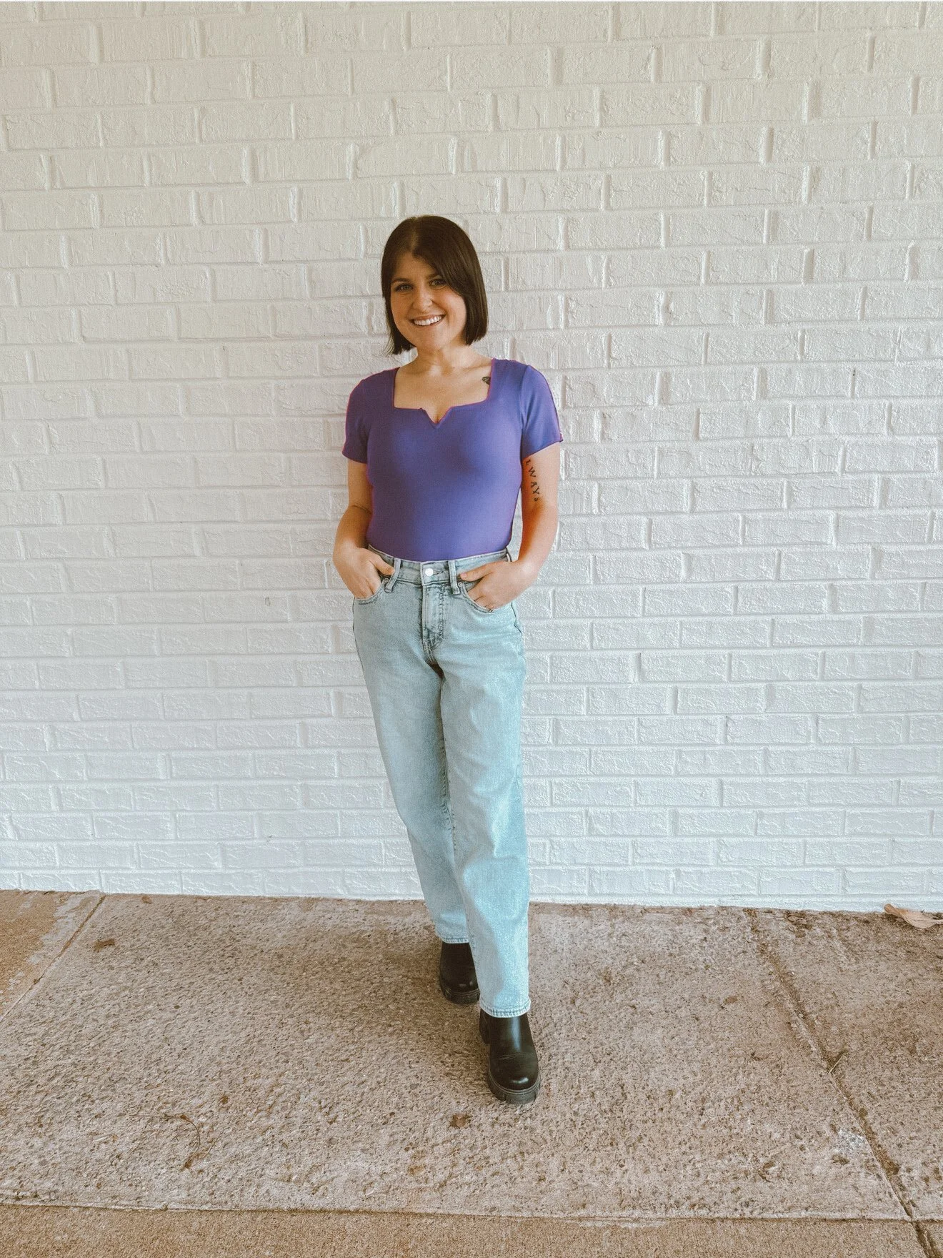 A woman with shoulder-length dark hair is standing in front of a white brick wall. She is smiling, wearing a purple short-sleeved top, light blue jeans, and black boots. Her hands are in her pockets.