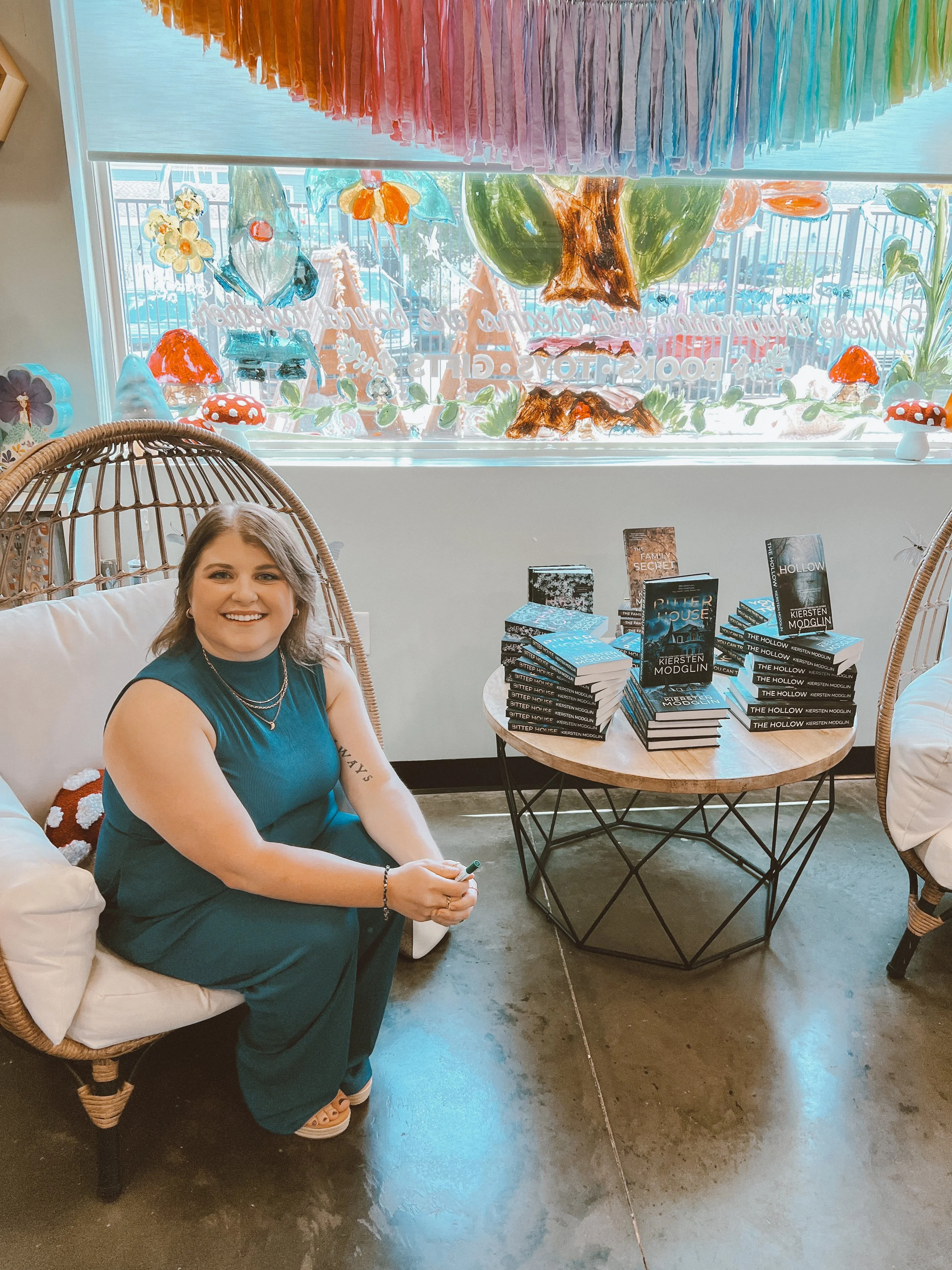 A woman sitting on a wicker chair in a bookstore or cafe, smiling, with a table of stacked books titled 'The Hollow' by Kiersten Modglin nearby, and colorful window decorations of painted nature scenes in the background.